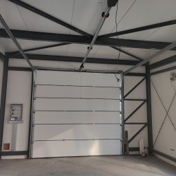 EIFAM Mateusz Nowacki - Wnętrze hali z zamontowaną segmentową bramą przemysłową w kolorze białym, widoczny mechanizm napędu pod sufitem oraz stalowa konstrukcja nośna budynku.