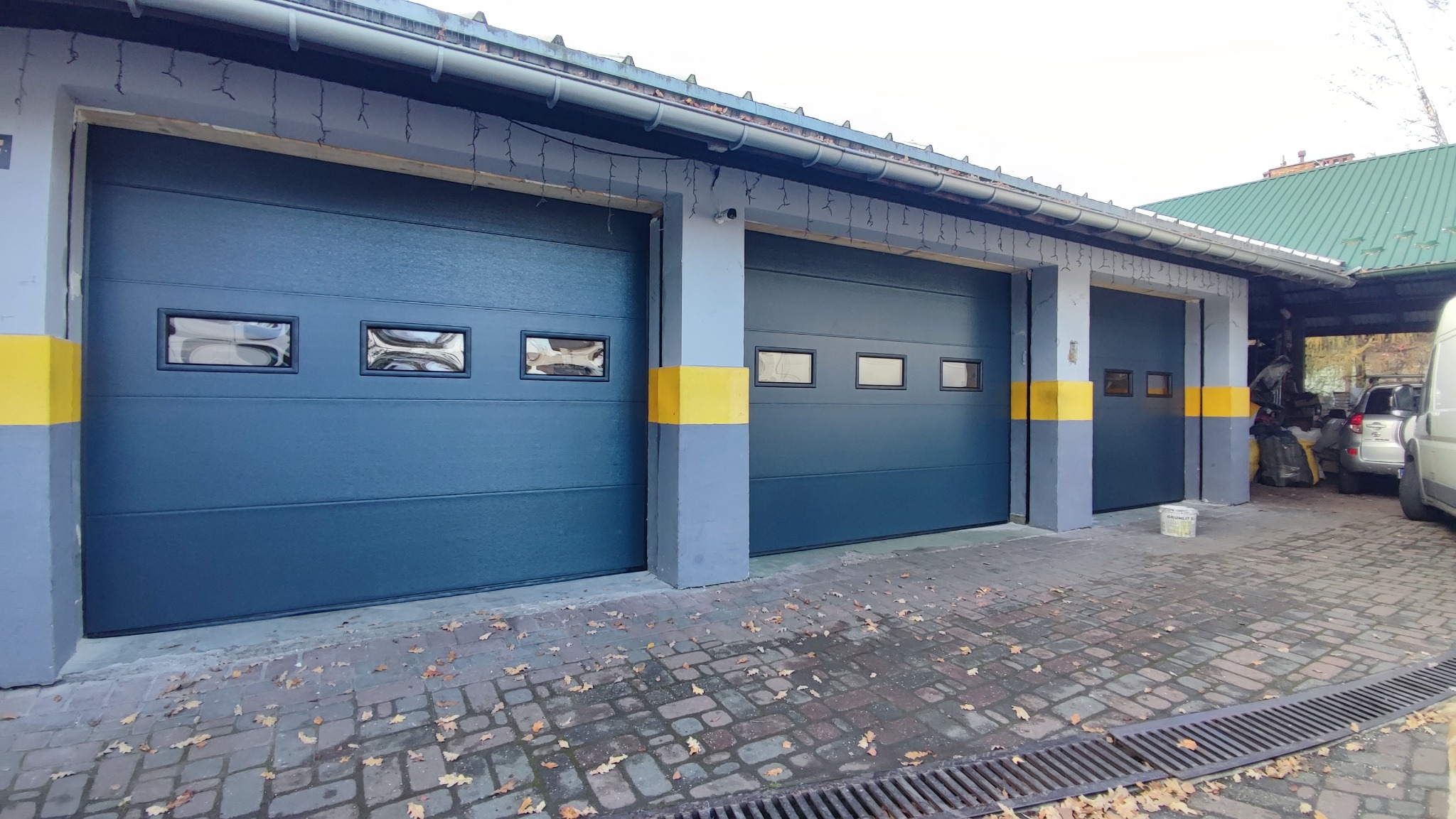 Trzy szare segmentowe drzwi z małymi okienkami w budynku z szarymi filarami z żółtymi akcentami, kostka brukowa pokryta liśćmi na zewnątrz.