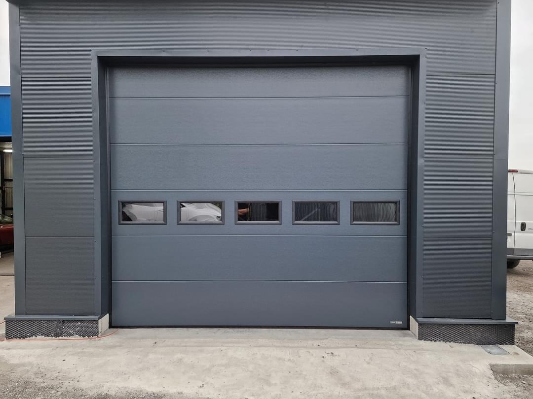 Szara brama garażowa segmentowa z poziomymi przetłoczeniami i rzędem małych, prostokątnych okienek, zamontowana w szarej elewacji budynku z blachy trapezowej.