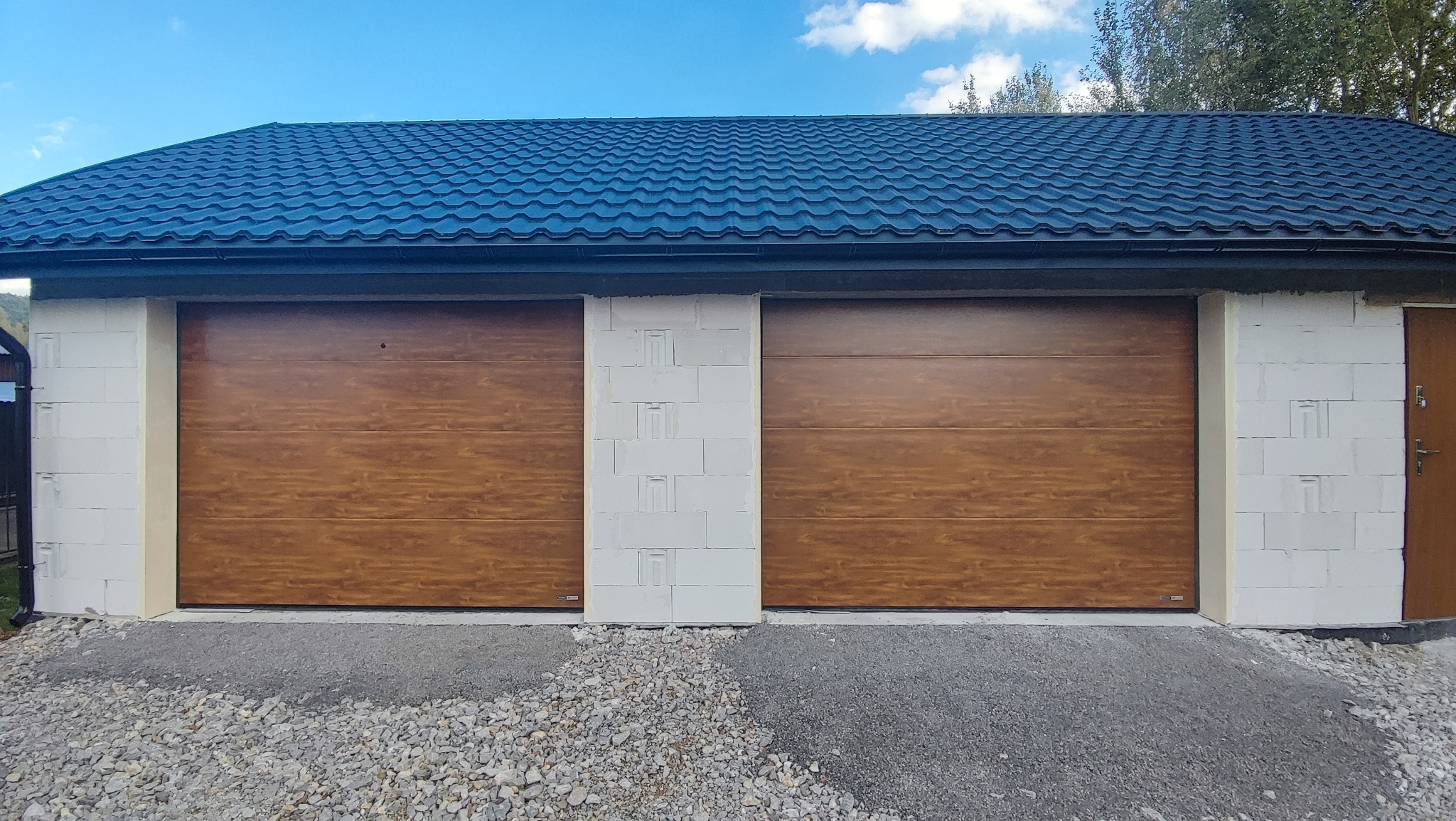 Dwie drewnopodobne, segmentowe bramy w garażu z białych bloczków, z ciemnym dachem z blachodachówki i żwirowym podjazdem.