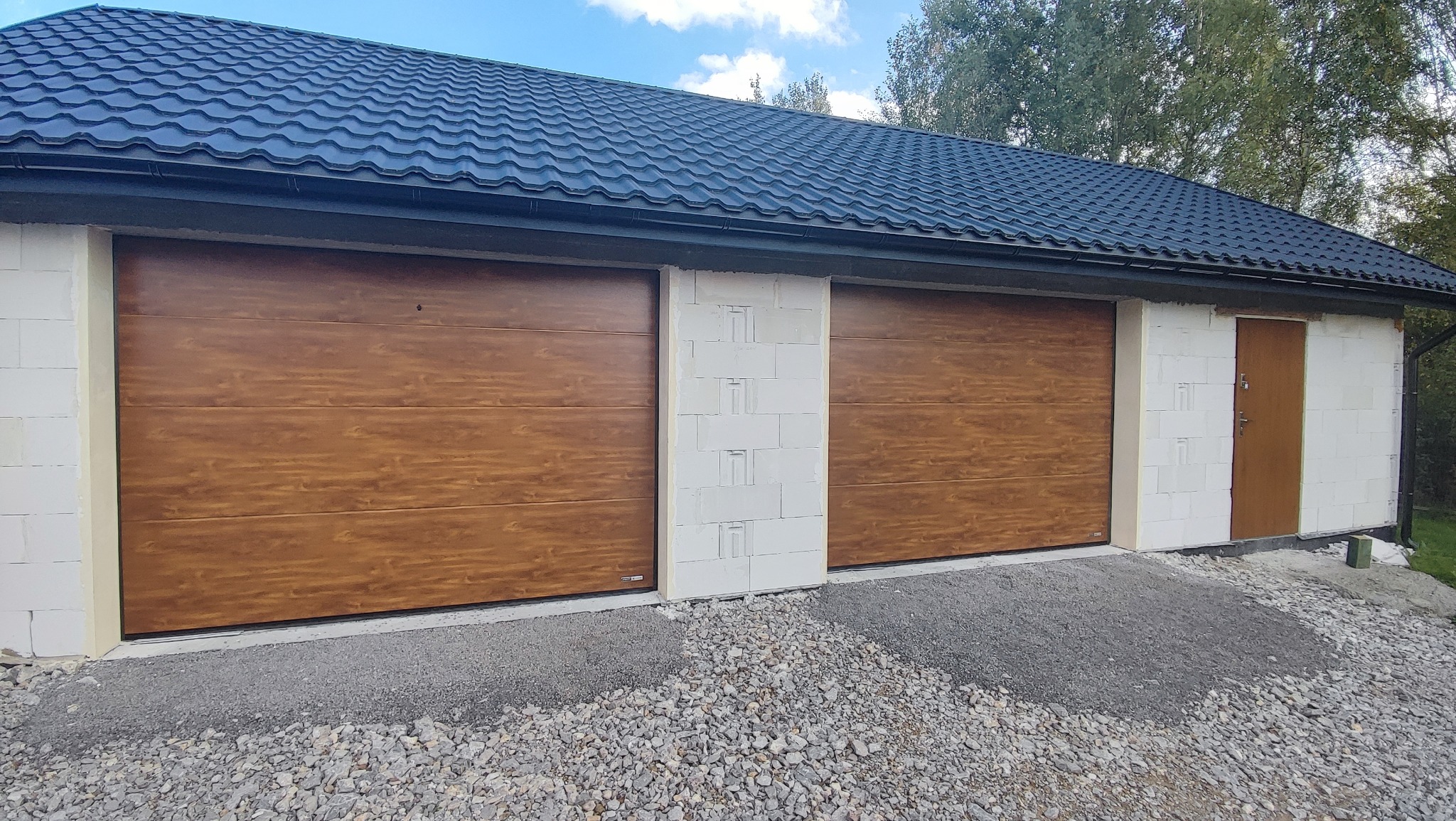 Nowoczesny garaż z dwoma drewnopodobnymi segmentowymi bramami, białe ściany z betonu komórkowego, czarny dach z blachodachówki, teren wokół wysypany żwirem.
