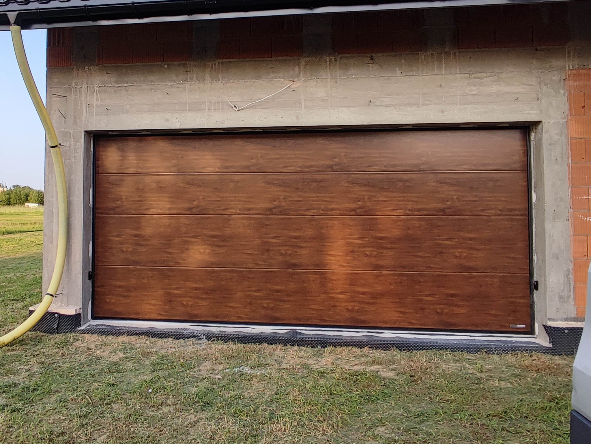 Zamknięta, segmentowa brama garażowa w kolorze drewna, zamontowana w surowym otworze budynku w trakcie budowy, z widoczną żółtą rurą wentylacyjną po lewej stronie i fragmentem trawnika u dołu.