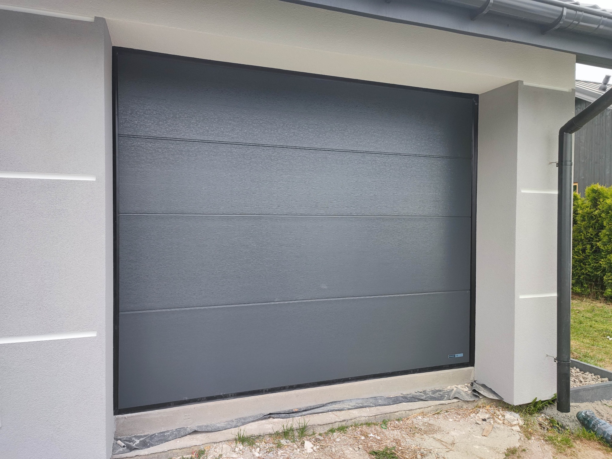 Zamknięta, segmentowa brama garażowa w kolorze grafitowym, z poziomymi przetłoczeniami, zamontowana w jasnoszarym budynku z widocznym systemem rynnowym.