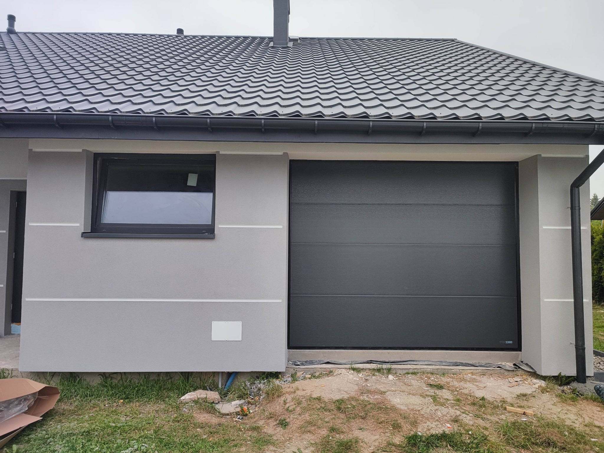 Nowoczesna, segmentowa, grafitowa brama garażowa w nowo wybudowanym domu z szarą elewacją i grafitowym dachem, widoczna z zewnątrz, z fragmentem trawnika i kartonem po prawej stronie.