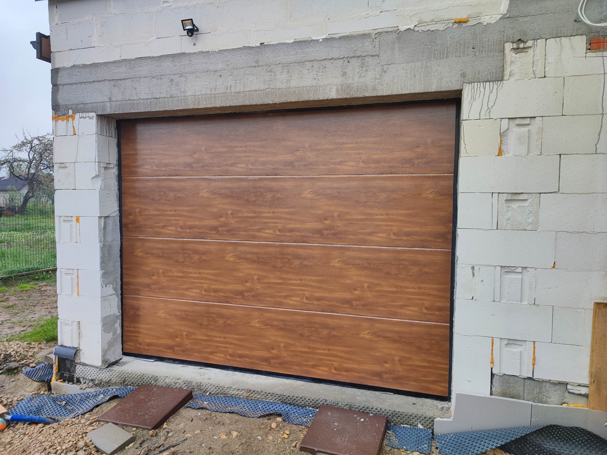 Nowa, segmentowa brama garażowa o drewnopodobnym wzorze, zamontowana w surowym stanie budynku w Nowym Sączu. Widoczne niedokończone obramowanie z białych bloczków.