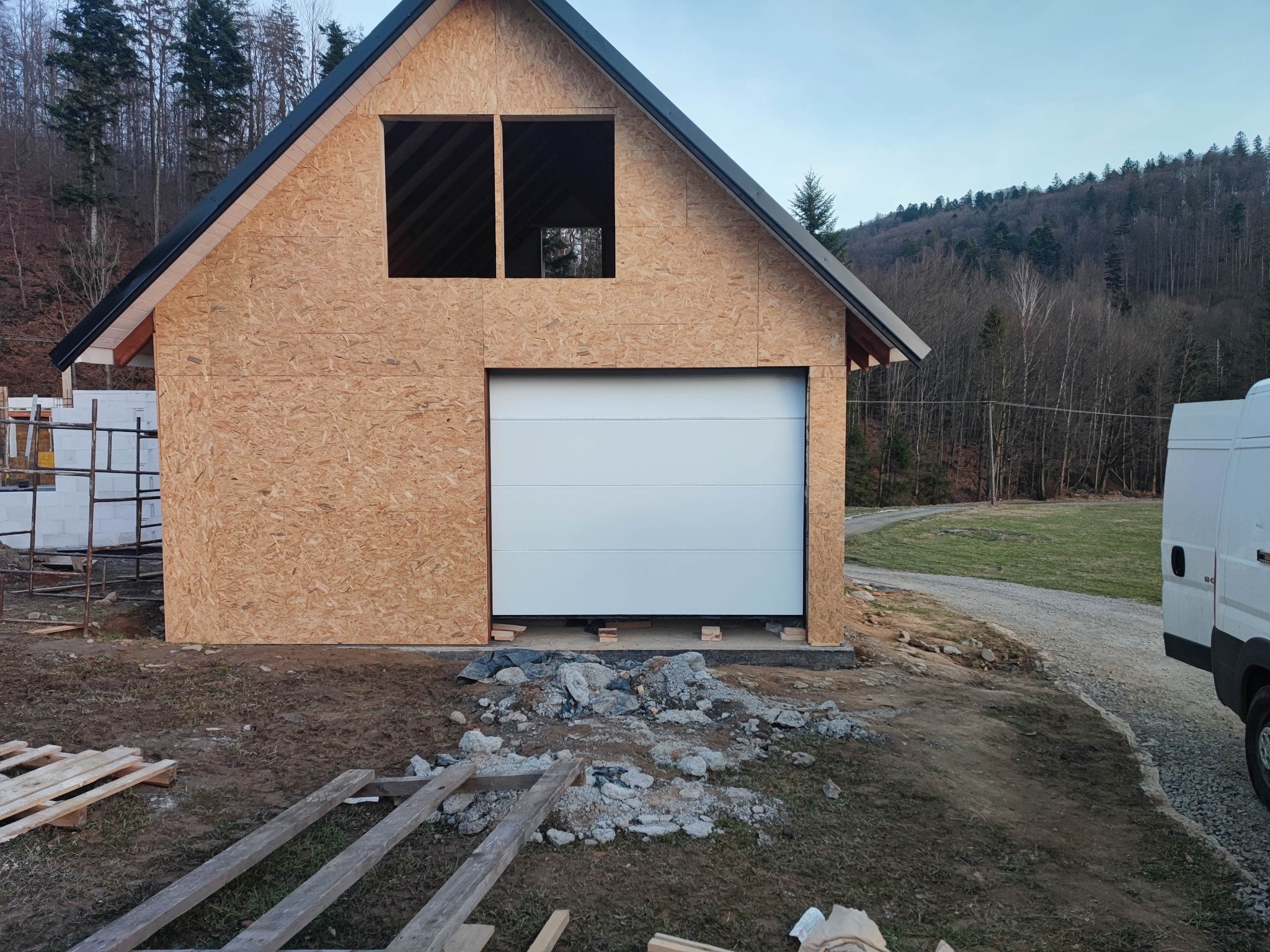 Nowa, biała brama segmentowa zamontowana w surowym garażu z płyty OSB, na zewnątrz gruz i nieporządek budowlany.