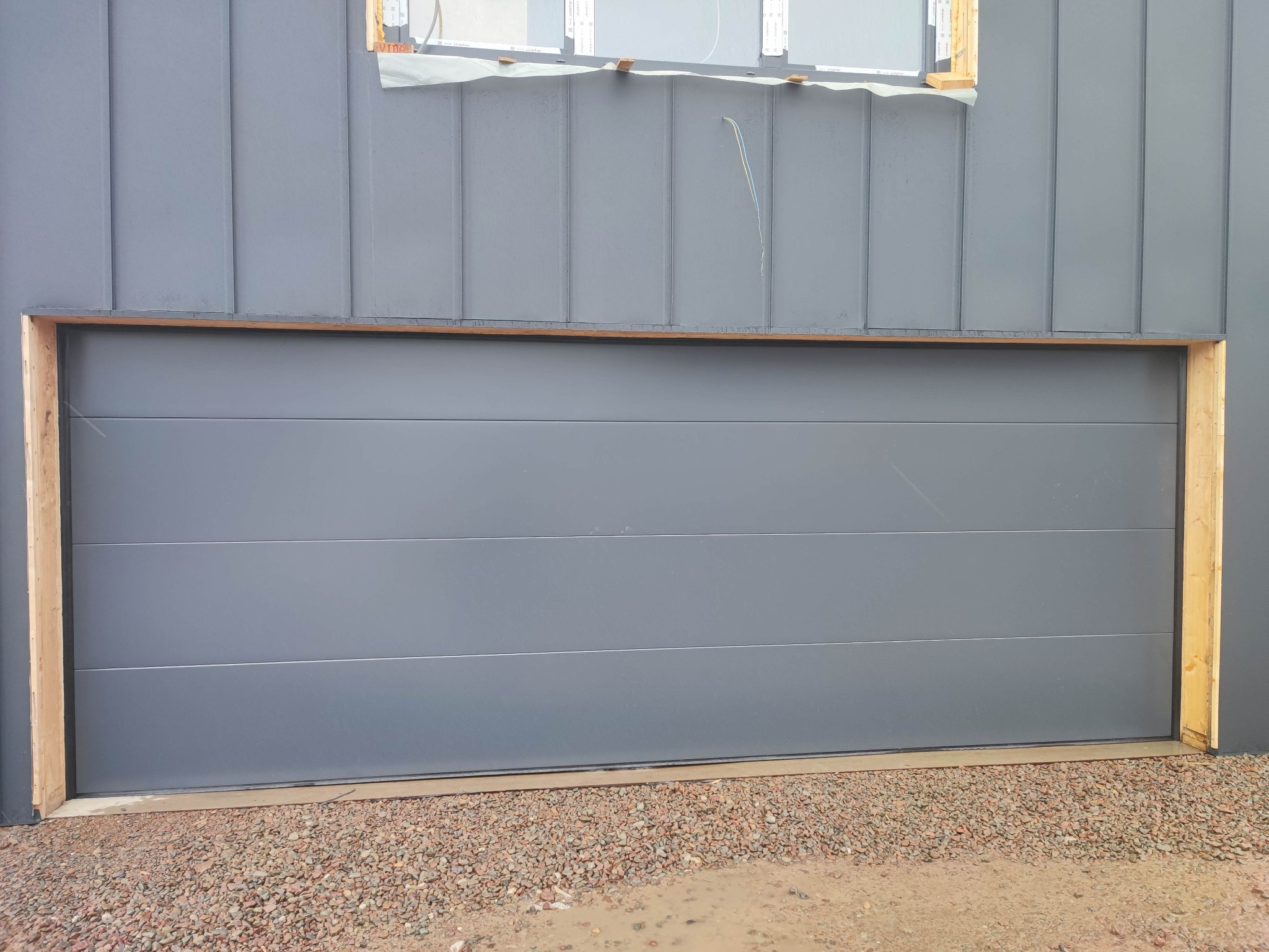 Szara brama segmentowa w trakcie montażu, widoczne drewniane ramy ościeżnicy, elewacja w pionowe panele i okno w stanie surowym.