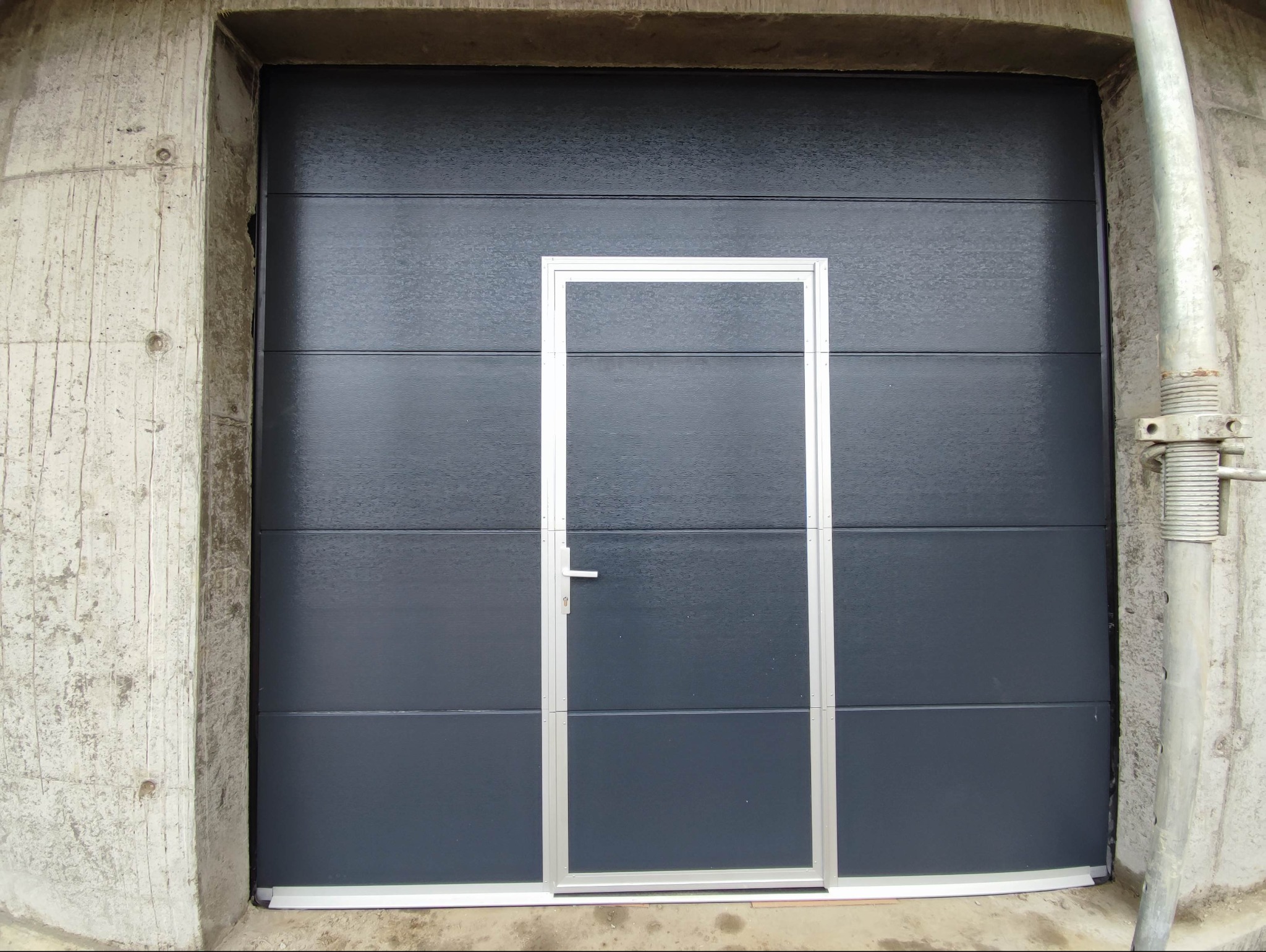 Grafitowa brama garażowa segmentowa z drzwiami serwisowymi z aluminiową ramą, zamontowana w surowym betonowym otworze, widoczna podpora budowlana.