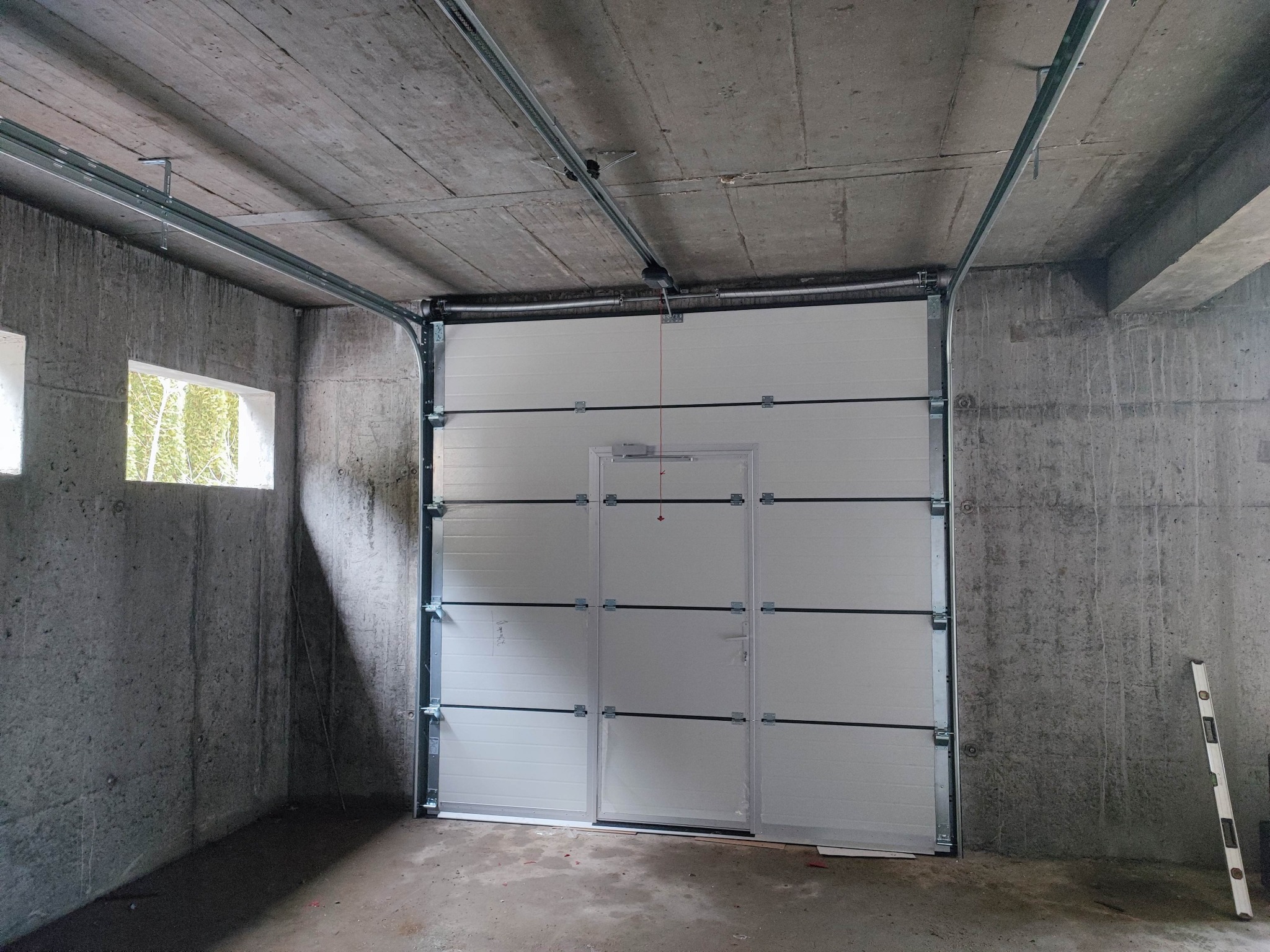 Zamknięta segmentowa brama garażowa z drzwiami przejściowymi w surowym, betonowym garażu; widoczna poziomica oparta o ścianę.