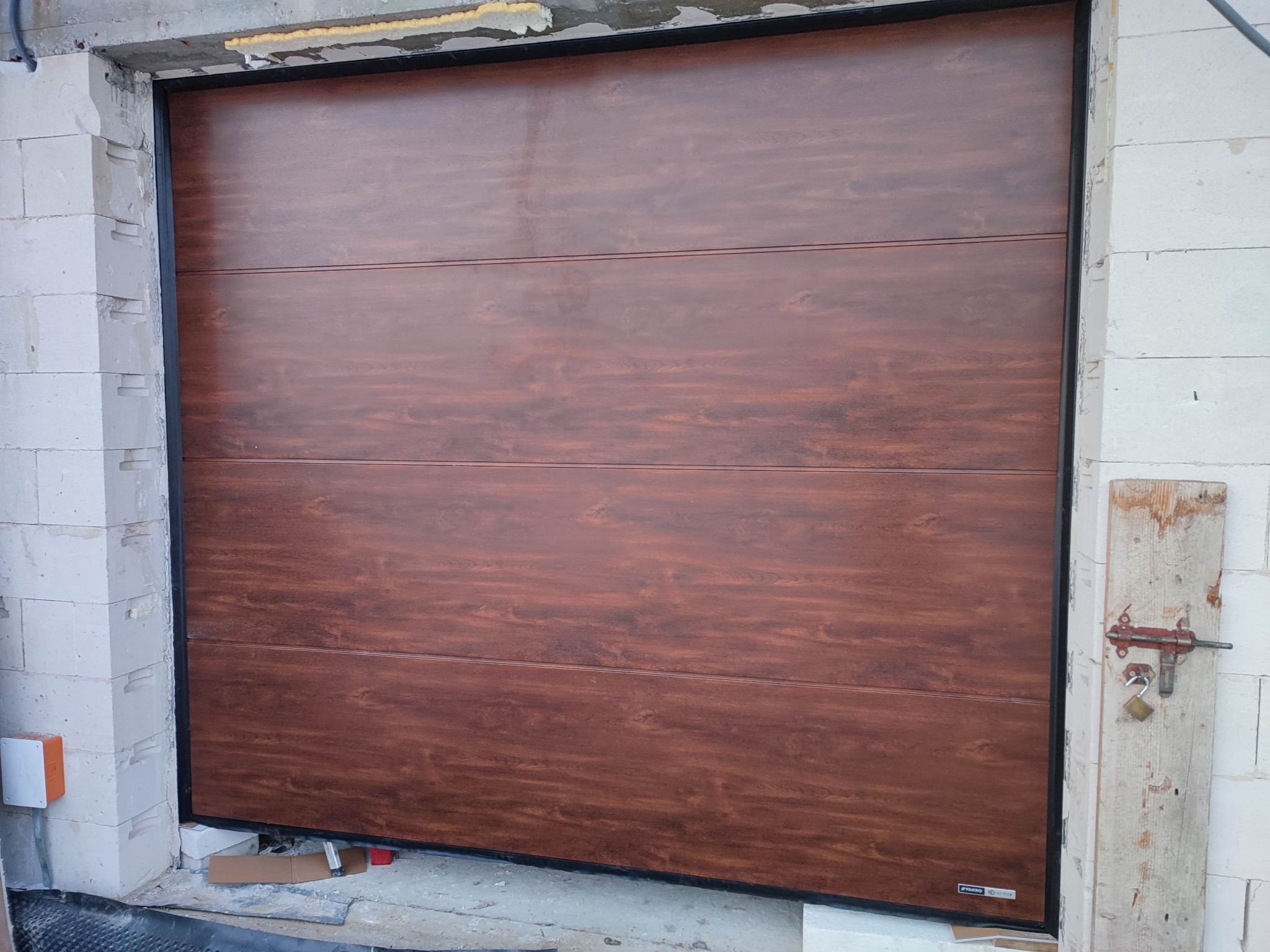 Zamknięta segmentowa brama garażowa w kolorze ciemnego drewna, widoczna od wewnątrz, z zamkiem na drewnianej listwie po prawej stronie i fragmentem surowej ściany z betonu komórkowego.
