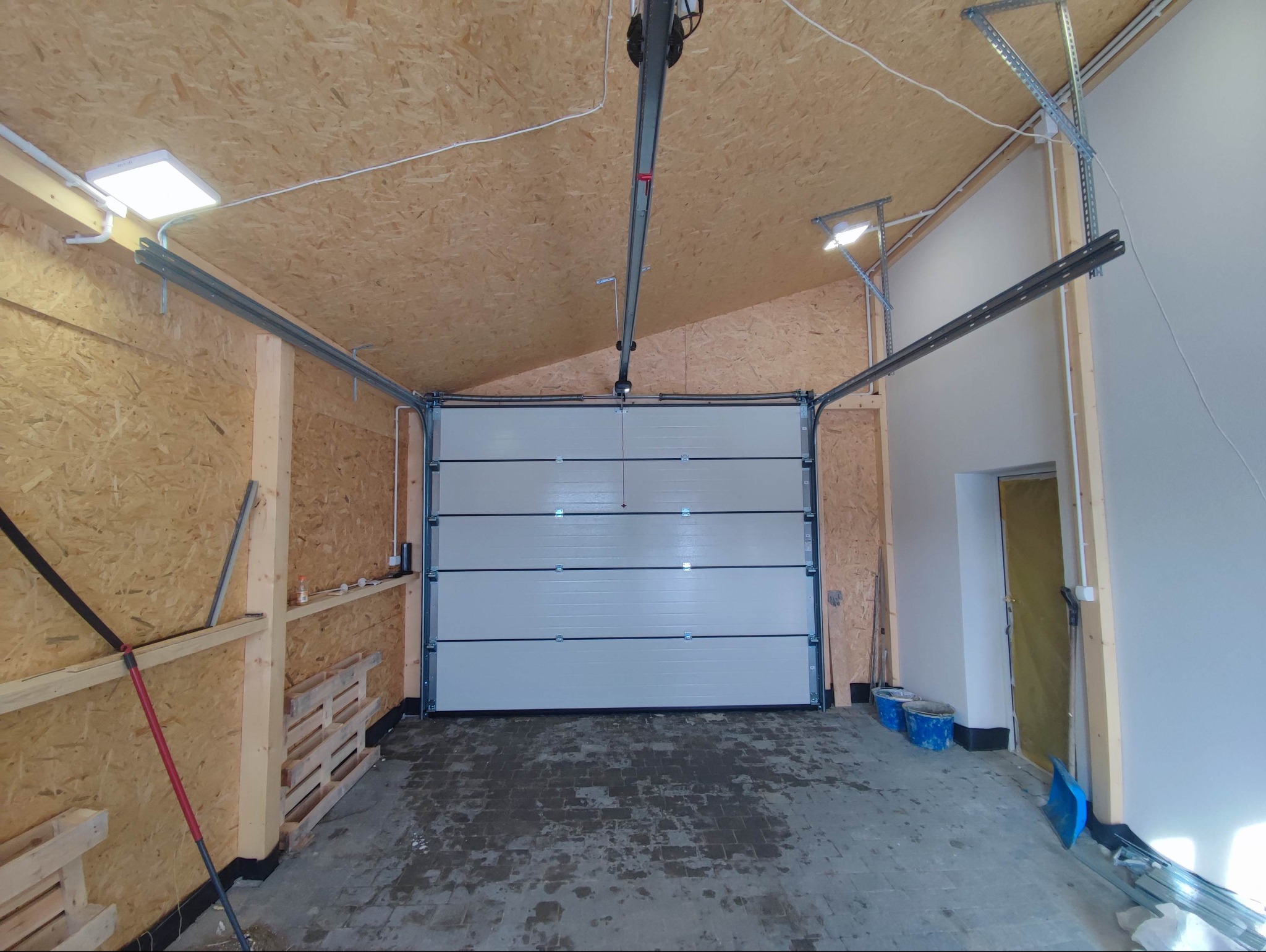 Nowa, segmentowa, biała brama garażowa zamontowana w garażu z drewnianym wykończeniem ścian i sufitu. Widoczne szyny prowadzące i mechanizm napędowy pod sufitem. Na podłodze ślady po pracach...