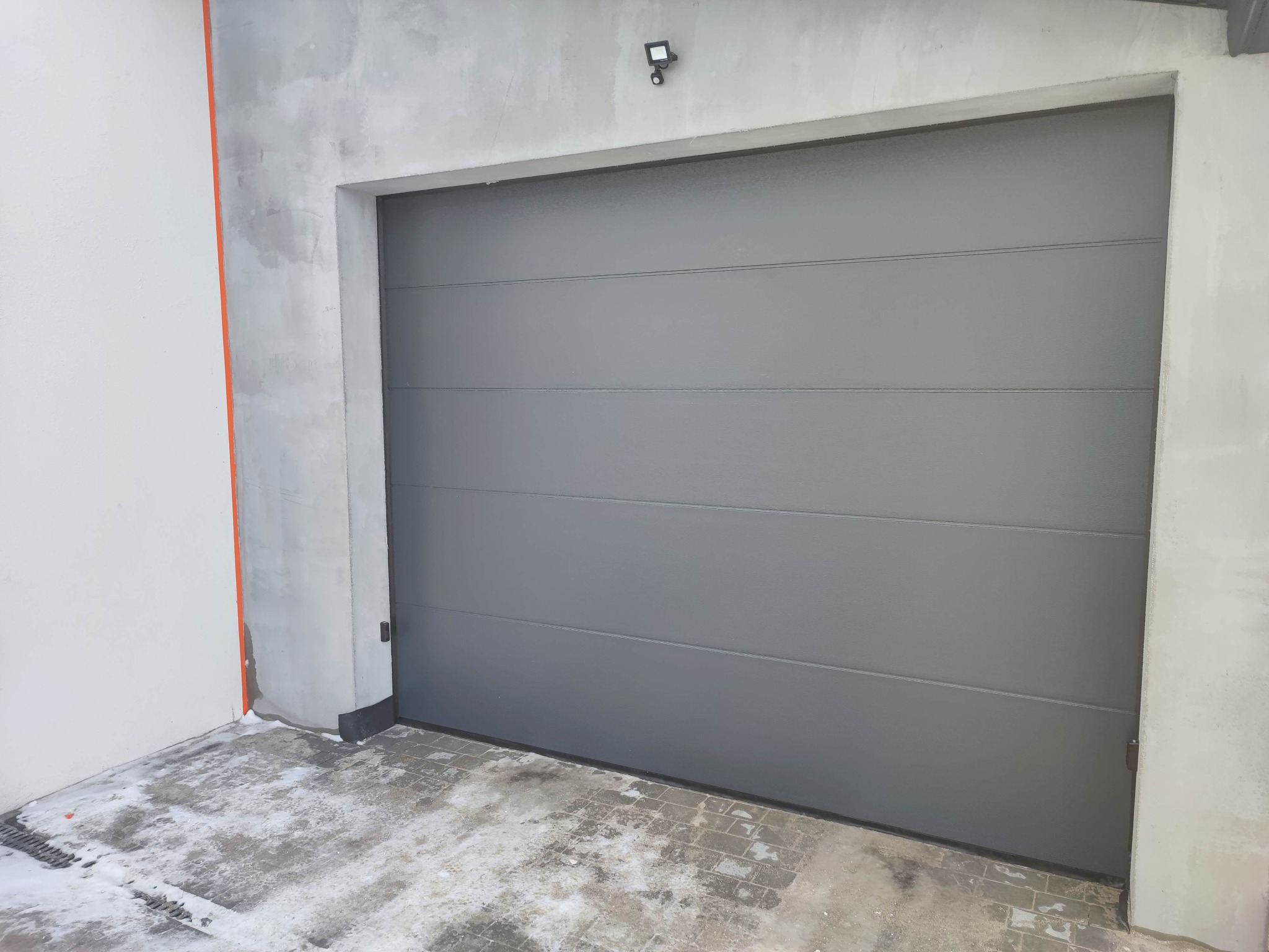 Szara segmentowa brama garażowa z poziomymi przetłoczeniami, zamontowana w surowym, otynkowanym otworze, z widocznym oświetleniem zewnętrznym i fragmentami śniegu na posadzce.