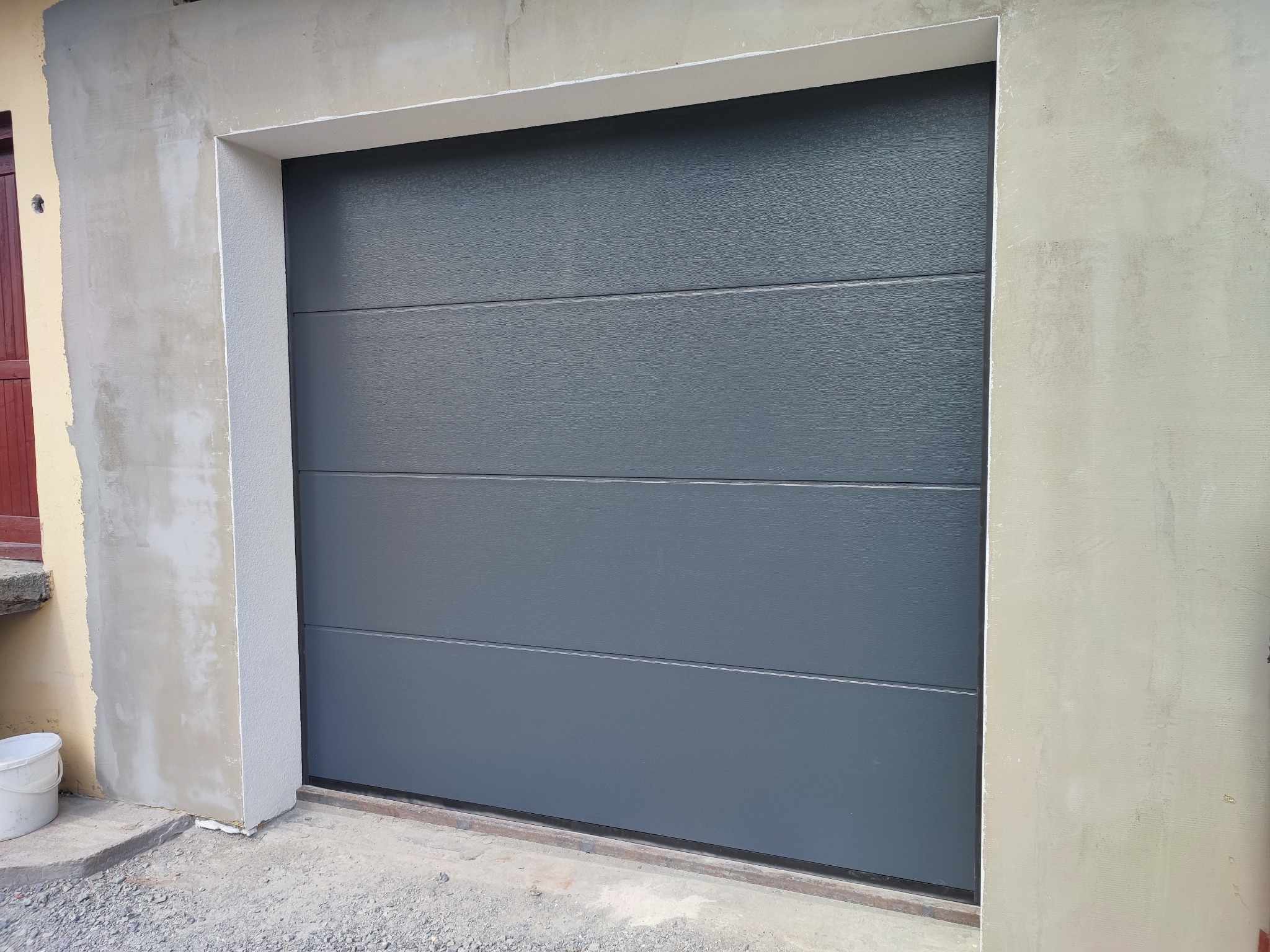 Zamknięta, segmentowa brama w kolorze grafitowym, z poziomymi przetłoczeniami, osadzona w surowej ramie z betonu i świeżego tynku.