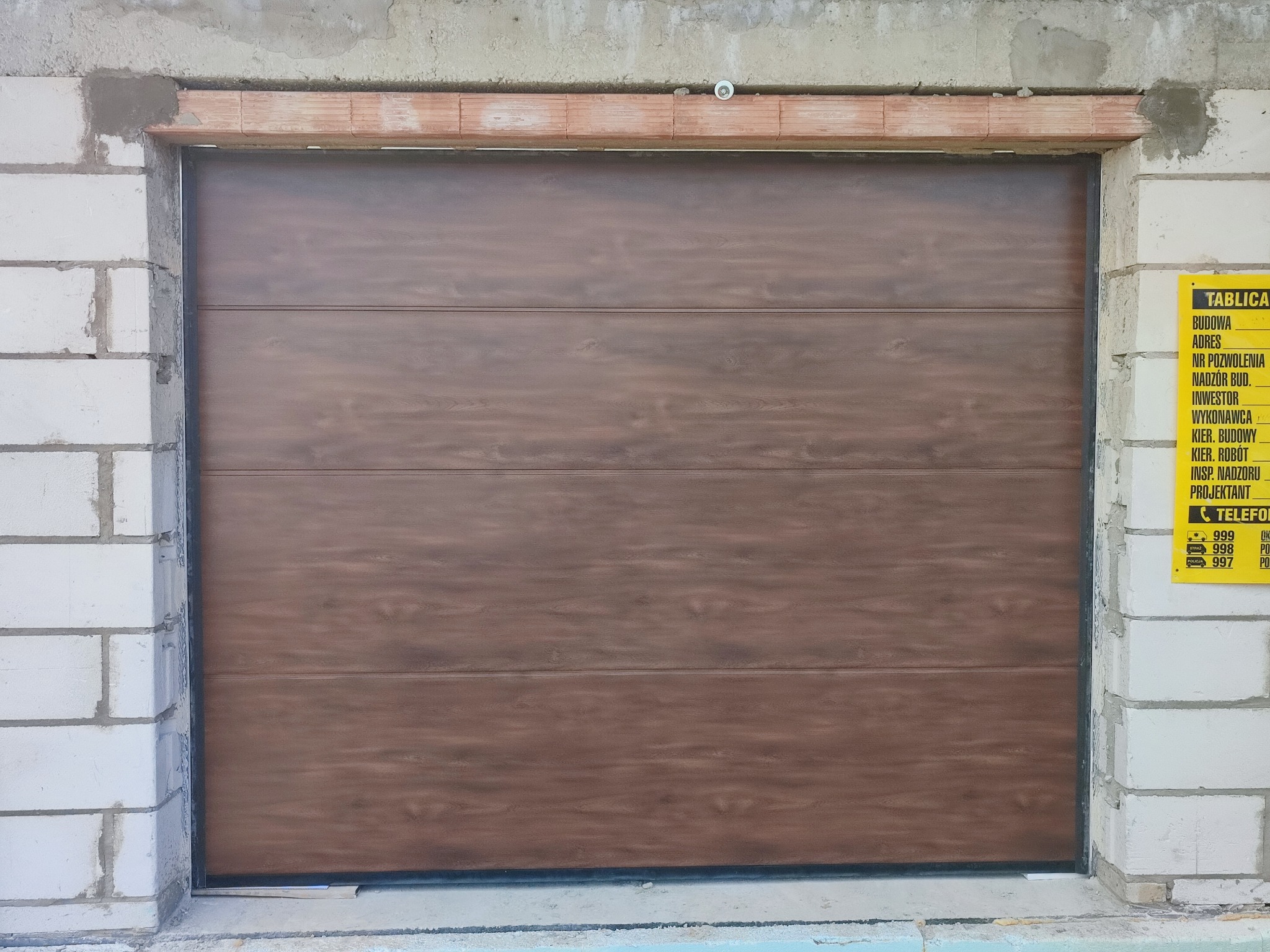 Zamknięta brama garażowa segmentowa w kolorze brązowego drewna, zamontowana w surowym otworze murowanym z bloczków betonowych, obok żółta tablica informacyjna budowy.