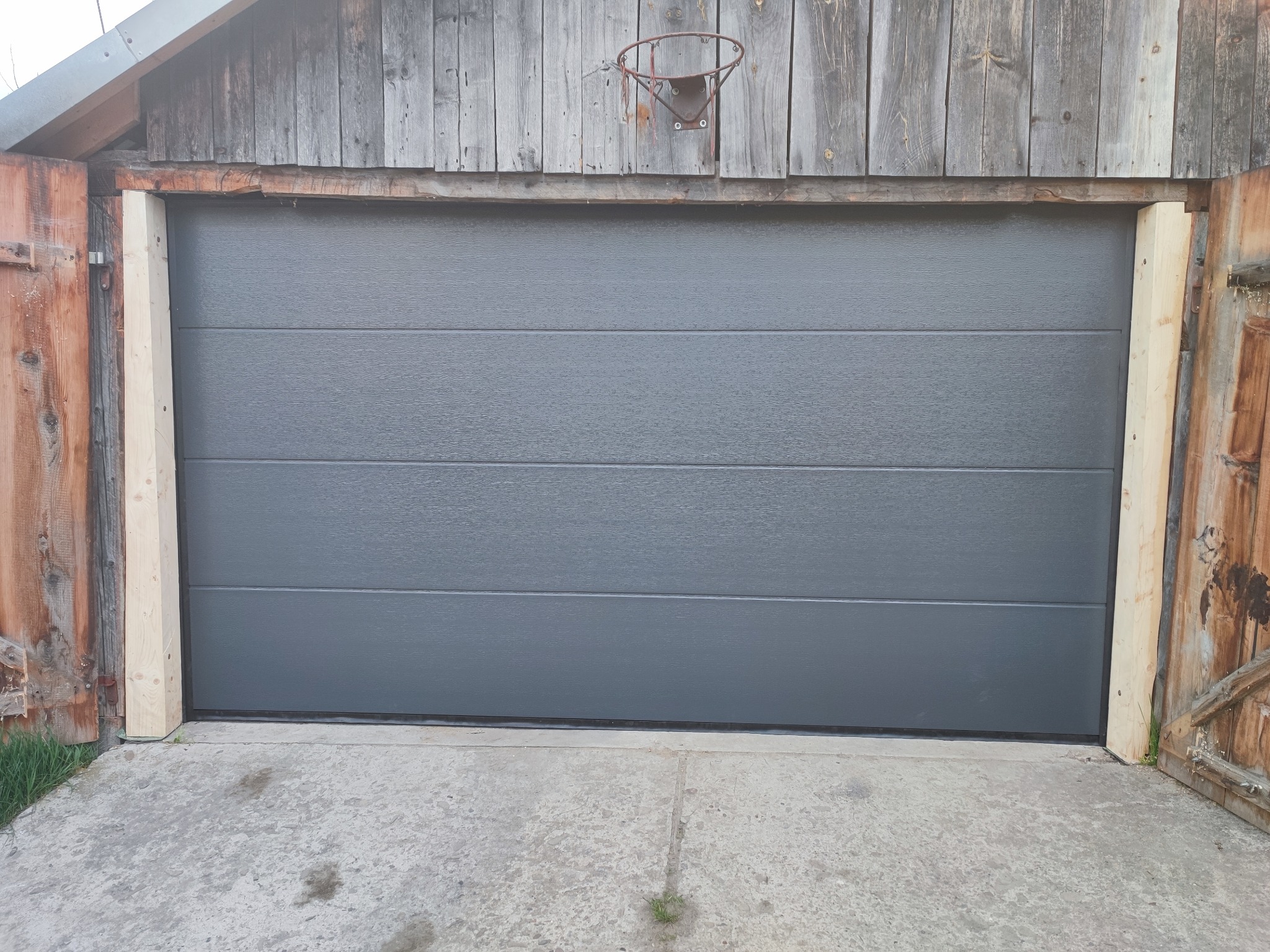 Szara segmentowa brama w stodole z drewnianą konstrukcją, widoczny stary kosz do koszykówki nad bramą.