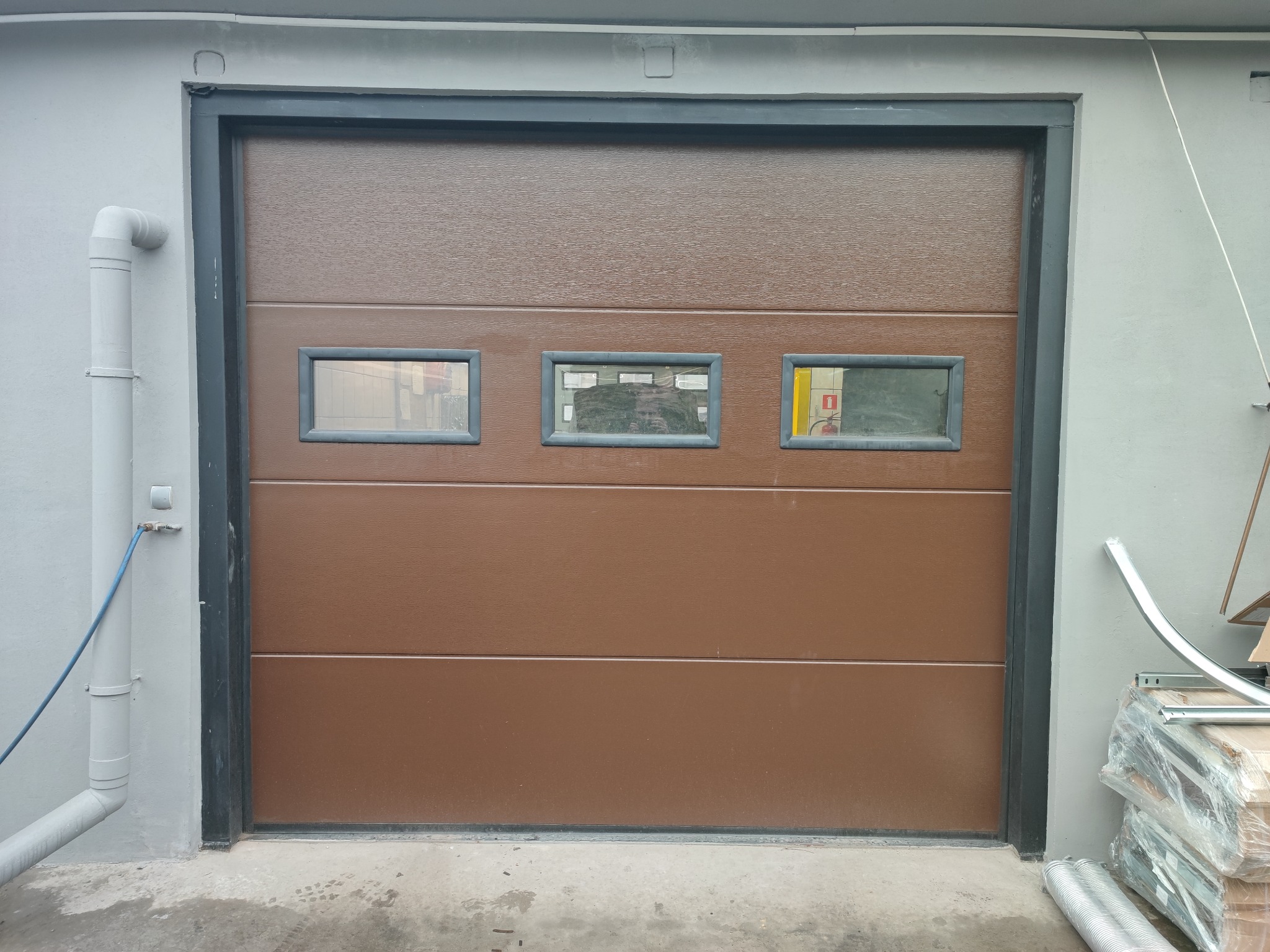 Brązowa segmentowa brama garażowa z trzema małymi okienkami, widoczna z zewnątrz, z szarą ramą i rurą spustową po lewej stronie, oraz materiałami budowlanymi po prawej stronie.