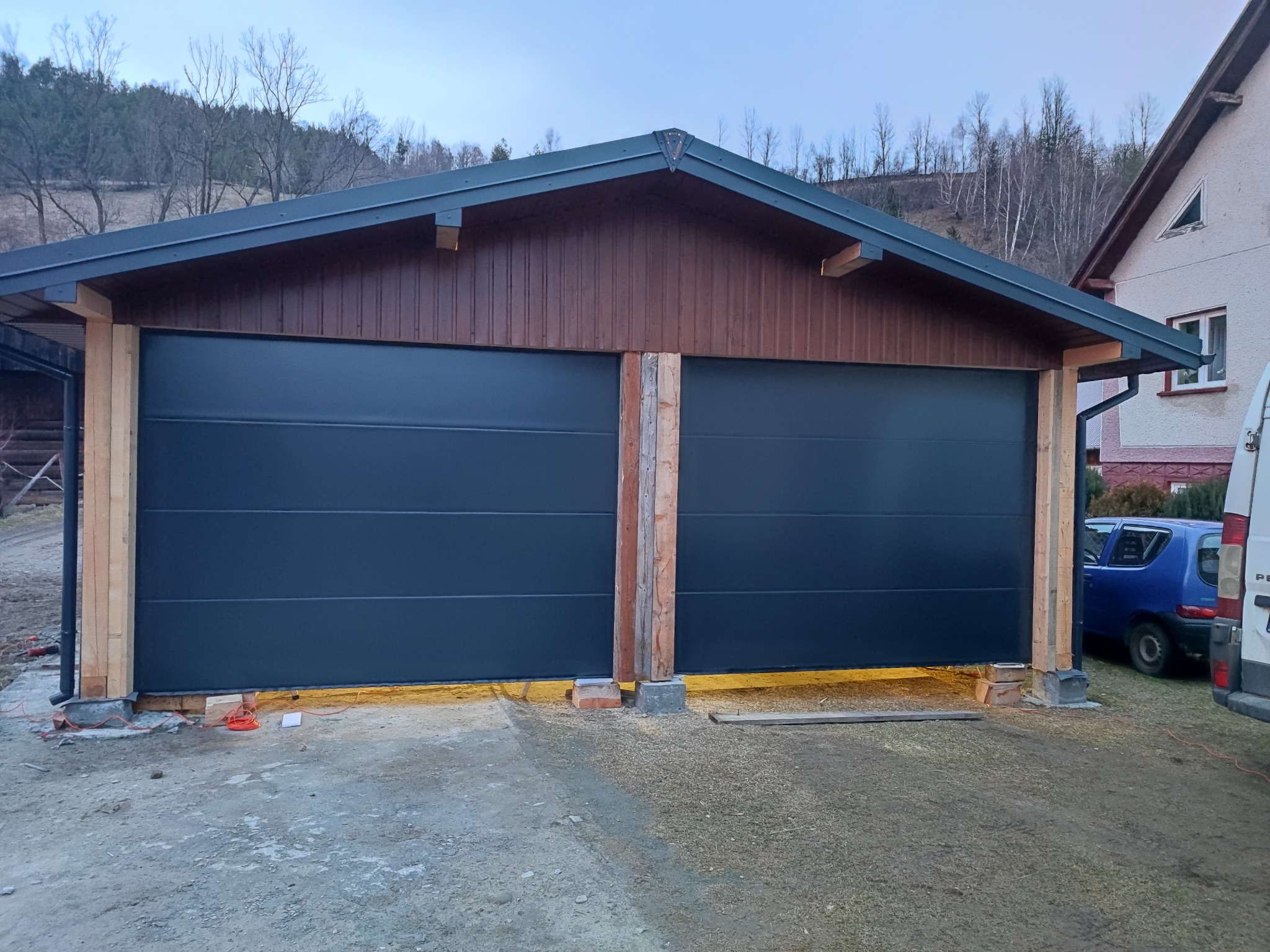 Dwustanowiskowy garaż z dwiema segmentowymi bramami w kolorze antracytowym, drewniana konstrukcja w trakcie montażu, widoczne przewody i cegły stabilizujące.