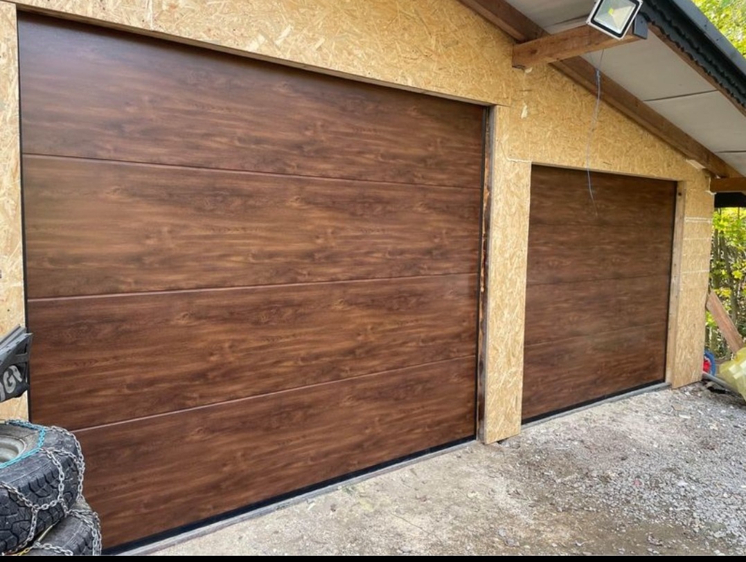 Dwie segmentowe bramy garażowe z efektem drewna, zamontowane w budynku z płyty OSB, widoczne opony zimowe z łańcuchami w rogu kadru.