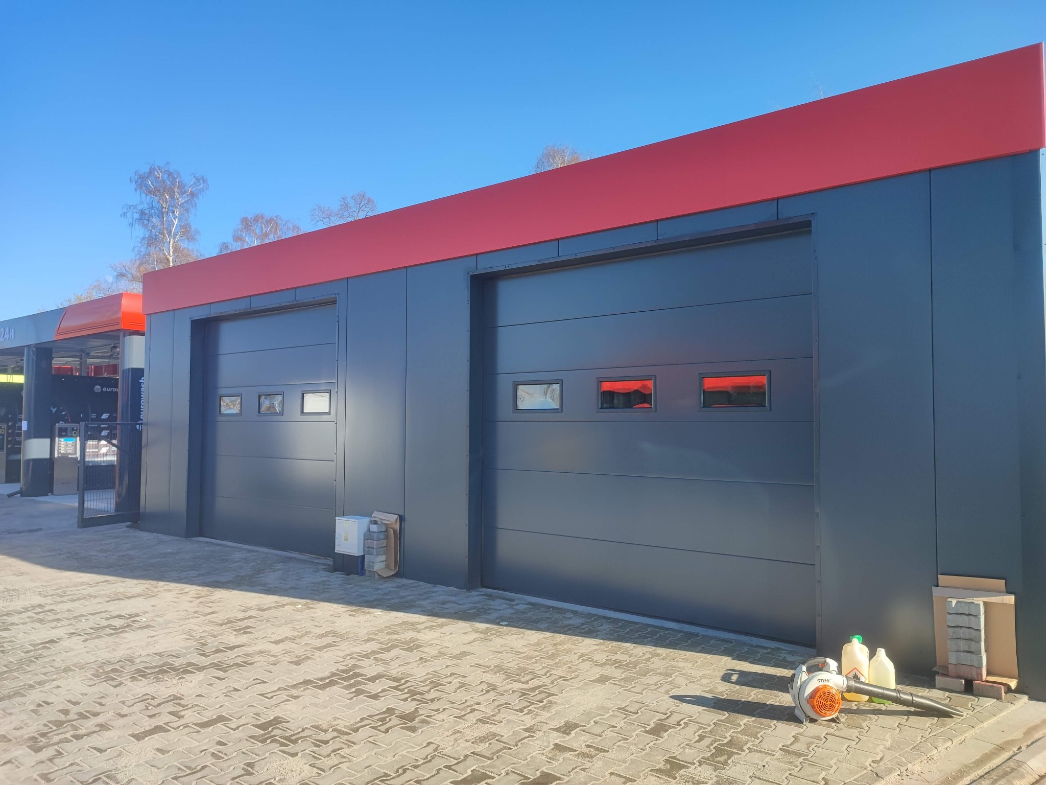 Dwa segmentowe wjazdy do hali przemysłowej w kolorze grafitowym z czerwonym pasem u góry, na kostce brukowej leży dmuchawa do liści i kanistry.