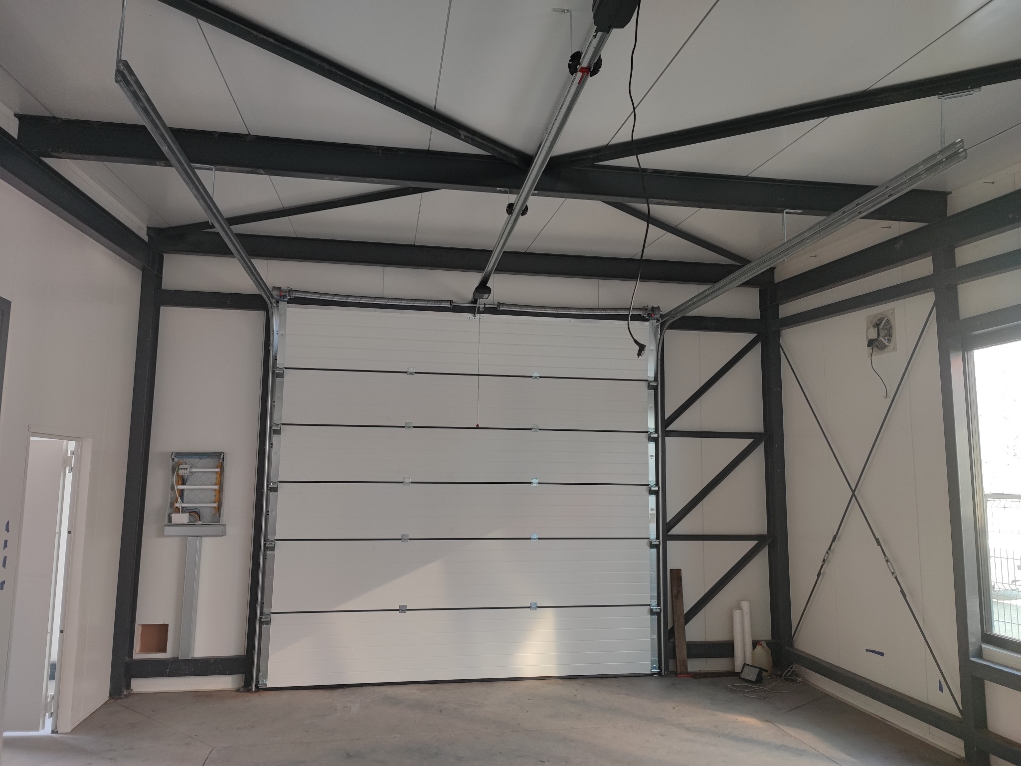 Wnętrze hali z zamontowaną segmentową bramą przemysłową w kolorze białym, widoczny mechanizm napędu pod sufitem oraz stalowa konstrukcja nośna budynku.