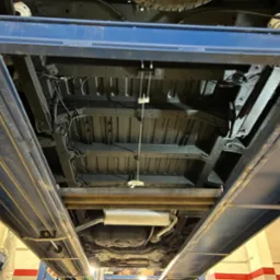 Podwozie samochodu po zabezpieczeniu antykorozyjnym, widoczne na podnośniku warsztatowym, z nową warstwą ochronną w kolorze czarnym, z widocznym układem wydechowym i elementami zawieszenia.