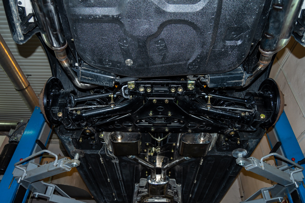 Podwozie samochodu po konserwacji antykorozyjnej, widoczne elementy zawieszenia, układ wydechowy i rama, podniesione na podnośniku warsztatowym.