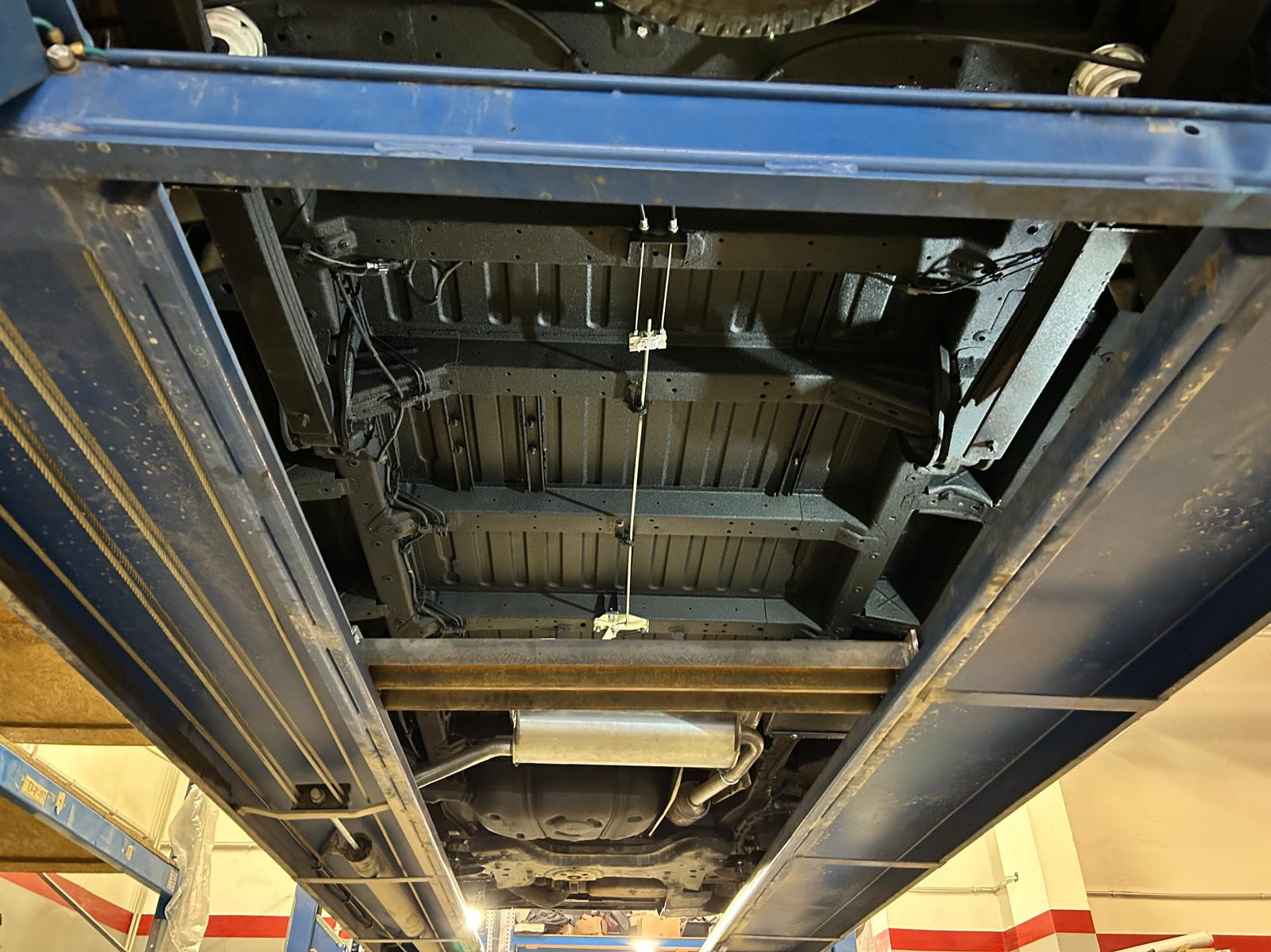 Podwozie samochodu po zabezpieczeniu antykorozyjnym, widoczne na podnośniku warsztatowym, z nową warstwą ochronną w kolorze czarnym, z widocznym układem wydechowym i elementami zawieszenia.