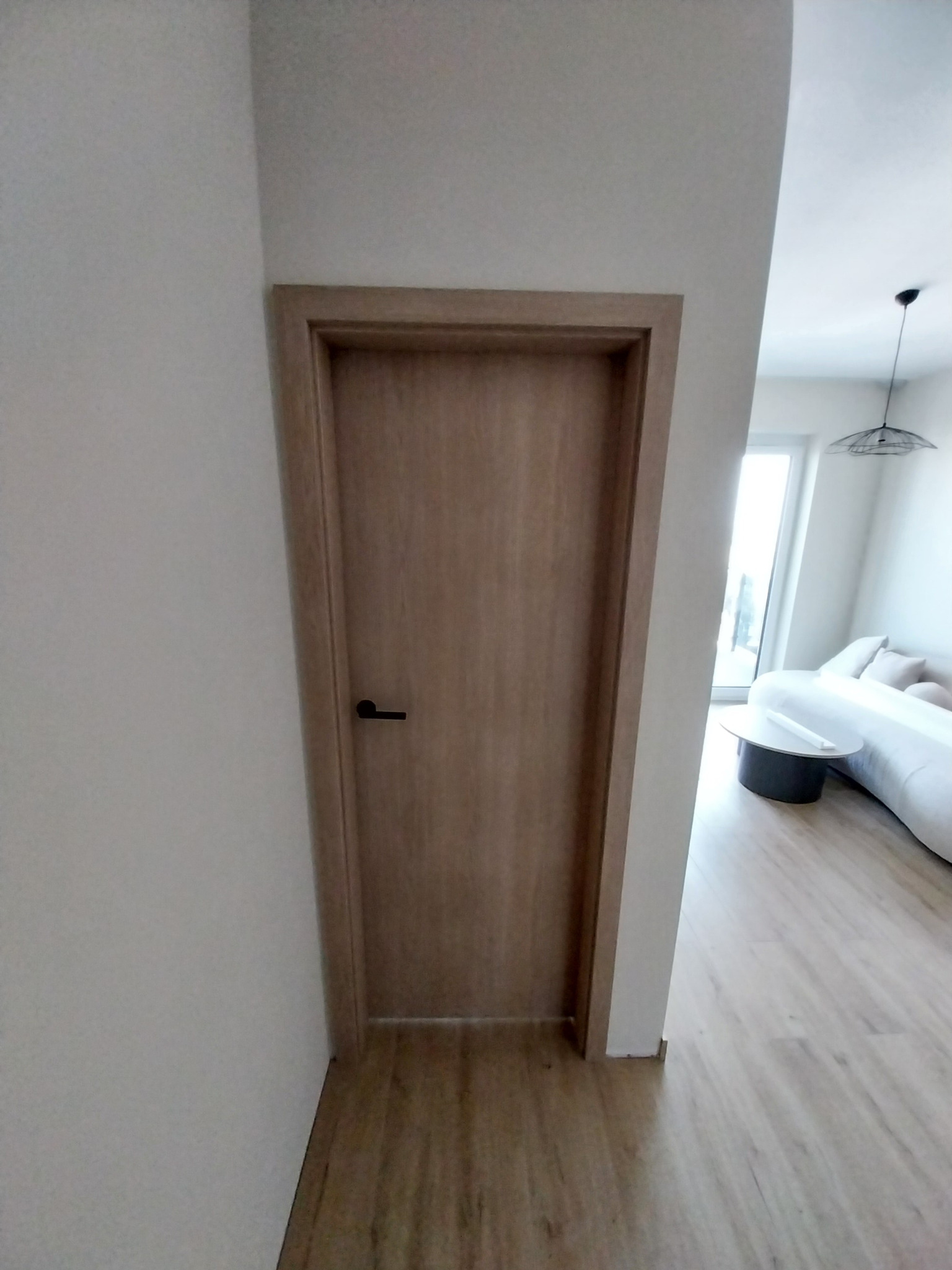 Zamknięte, drewniane drzwi wewnętrzne z czarną klamką, widoczne na tle jasnego wnętrza z parkietem i białymi meblami. Minimalistyczny design.