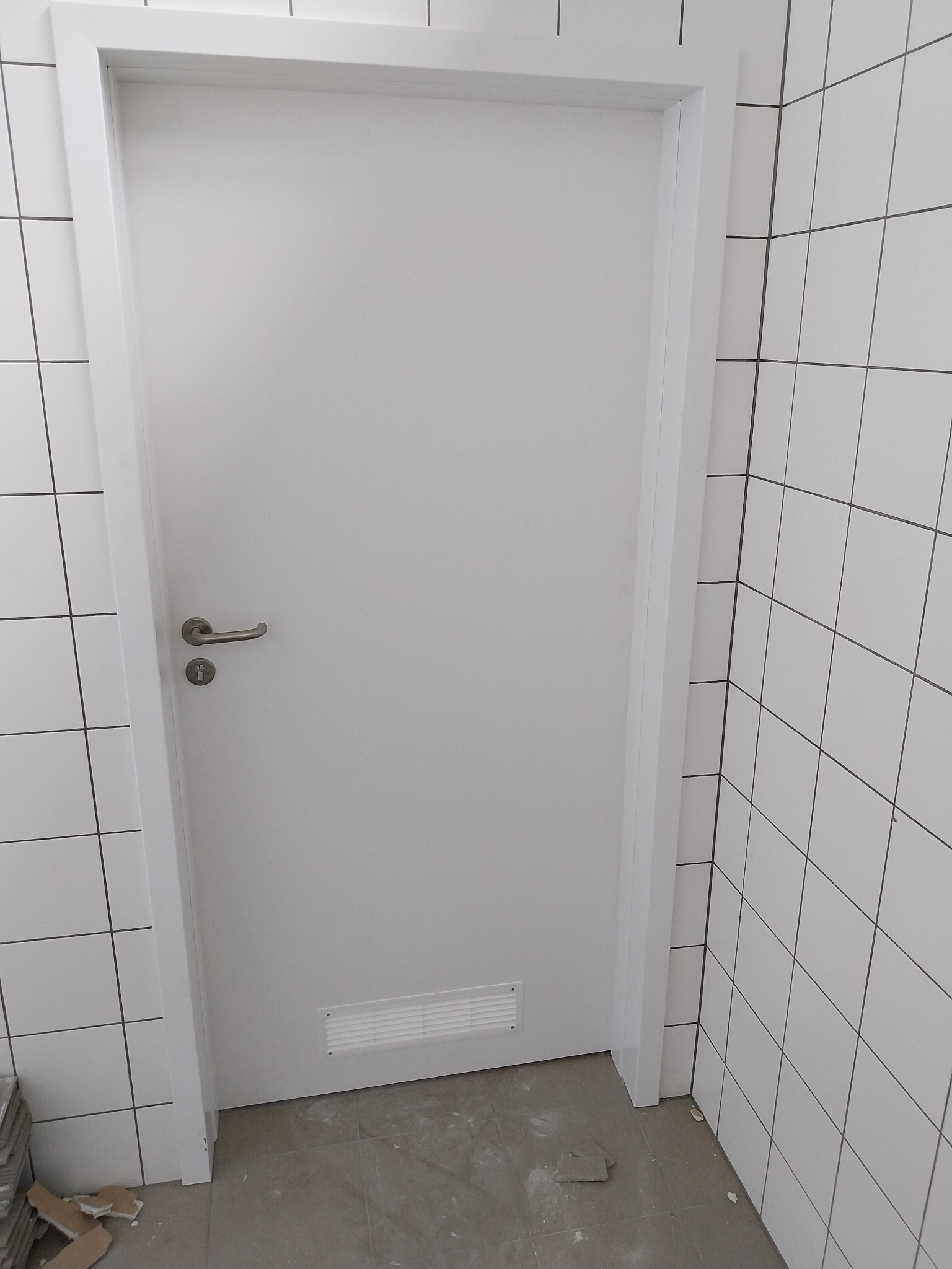 Białe, nowoczesne drzwi z wentylacją w dolnej części, zamontowane w łazience wykończonej białymi, kwadratowymi płytkami. Widoczna srebrna klamka i fragment podłogi.