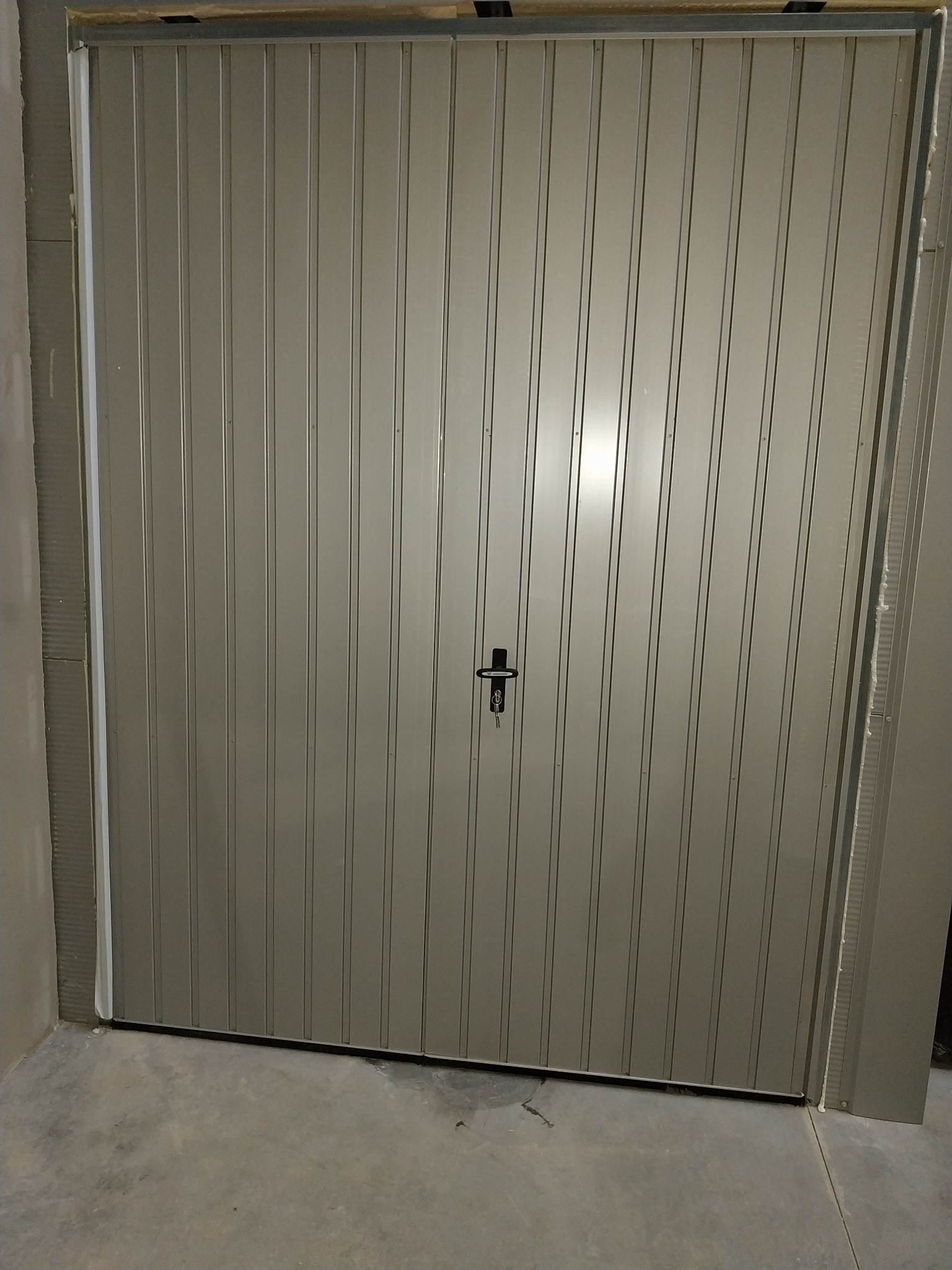 Szara, metalowa brama garażowa z pionowymi przetłoczeniami i czarnym zamkiem pośrodku, zamontowana w surowym, betonowym otoczeniu.