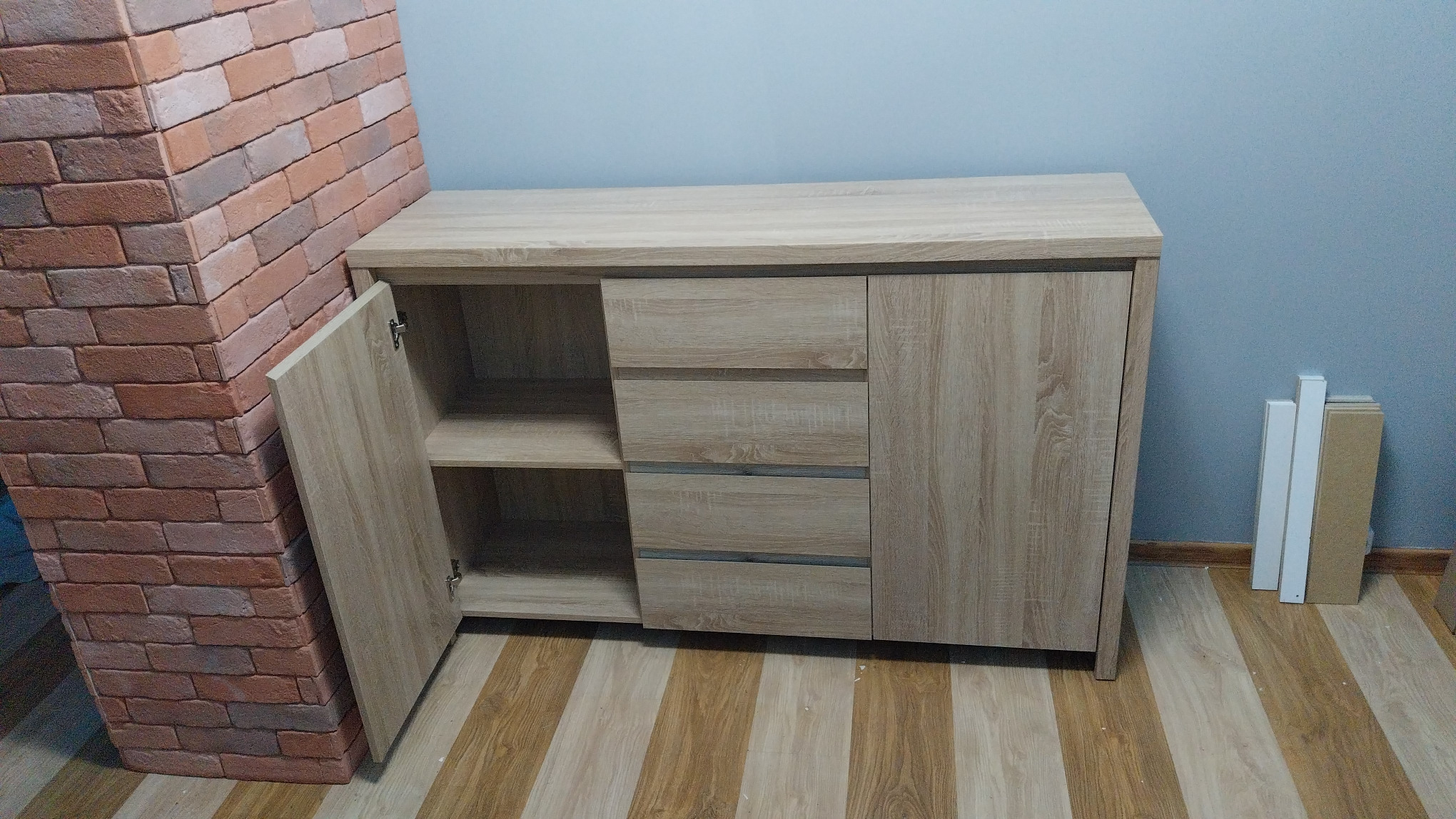 Beżowa komoda z otwartymi drzwiczkami, trzema szufladami i szafką, ustawiona przy ceglanej ścianie i szarej ścianie, na podłodze z paneli.