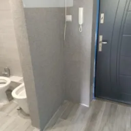 Szara ściana z domofonem i drzwiami wejściowymi w tle, widoczna toaleta i płytki podłogowe imitujące drewno. Wykończenie wnętrza w odcieniach szarości.