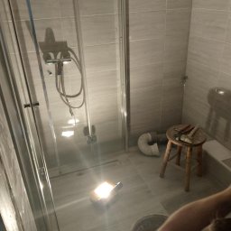 Robert Szczęsny - Szara łazienka w trakcie remontu: kabina prysznicowa, płytki, narzędzia na stołku, oświetlenie robocze. Widoczny fragment osoby. Ujęcie z bliska, podkreślające etap prac.