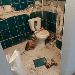 Ogólno budowlana Robert Szczęsny - Demontaż płytek w łazience z widocznym uszkodzeniem ściany, narzędziami i workiem na gruz. Widoczny etap prac remontowych z użyciem wiertarki i młota udarowego.