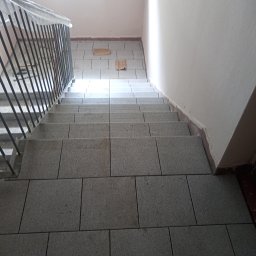 Ogólno budowlana Robert Szczęsny - Wykończone schody wewnątrz budynku, wyłożone szarymi, kwadratowymi płytkami. Metalowa barierka zabezpieczona folią. Widoczne ślady po pracach wykończeniowych.