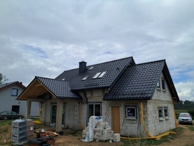 Dom jednorodzinny w trakcie budowy z czarnym dachem i widocznymi elementami konstrukcyjnymi. Na pierwszym planie materiały budowlane i przewody wentylacyjne.