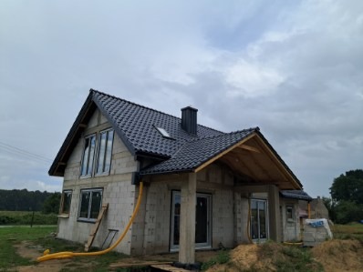 Dom jednorodzinny w trakcie budowy z czarnym dachem z dachówki, widoczne okna i rynny. Na pierwszym planie żółty wąż. Pochmurne niebo w tle.