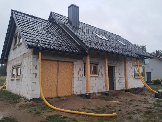 Dom w trakcie budowy z czarnym dachem i rynnami. Widoczne okna, komin i żółte rury wentylacyjne. Ściany z białego pustaka.