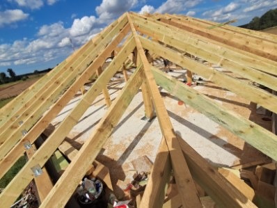 Konstrukcja drewniana dachu w trakcie budowy, widok z góry. Elementy złączone metalowymi płytkami, w tle błękitne niebo z chmurami i zieleń drzew.