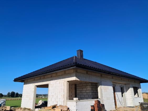 Surowy dom jednorodzinny w trakcie budowy, z czarnym dachem i kominem na tle błękitnego nieba. Widoczne pustaki i drewniane elementy konstrukcyjne.