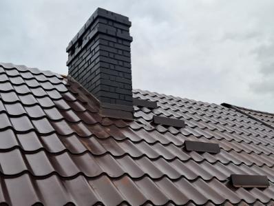 Ciemnobrązowy dach z blachy trapezowej z ceglanym kominem w kolorze grafitowym, zabezpieczony stopniami kominiarskimi. Pochmurne niebo w tle.