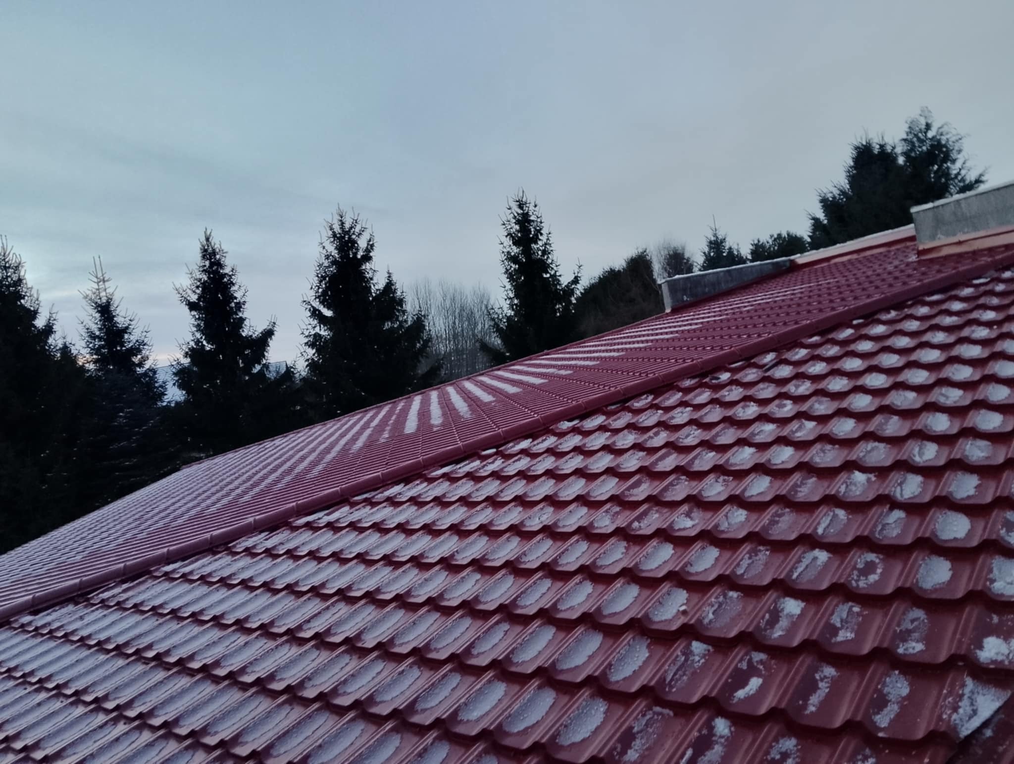 Czerwony dach z blachodachówki, pokryty lekkim śniegiem, na tle ciemnych drzew iglastych i pochmurnego nieba. Widok z góry, ukośny kadr.