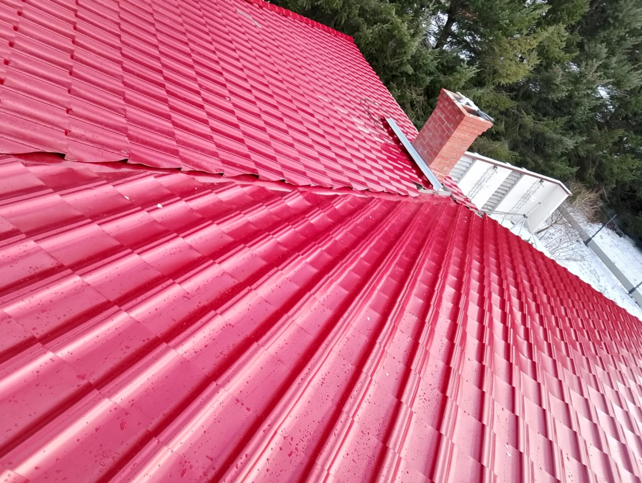 Czerwony dach z blachy trapezowej, widoczny komin i fragmenty drzew. Świeżo położone pokrycie dachowe w słoneczny dzień.