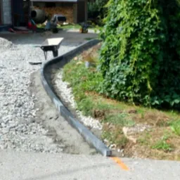 Krawężnik z betonowych elementów oddziela obszar z szarą kostką od trawnika z zieloną roślinnością, w tle widoczna taczka i betoniarka.