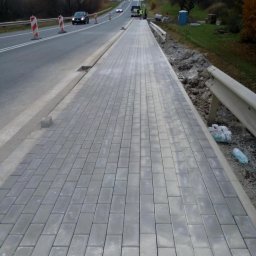 Malwina Zajdel - Świeżo ułożona, szara kostka brukowa tworząca chodnik wzdłuż drogi, z widocznymi samochodami w oddali i elementami zabezpieczającymi budowę.