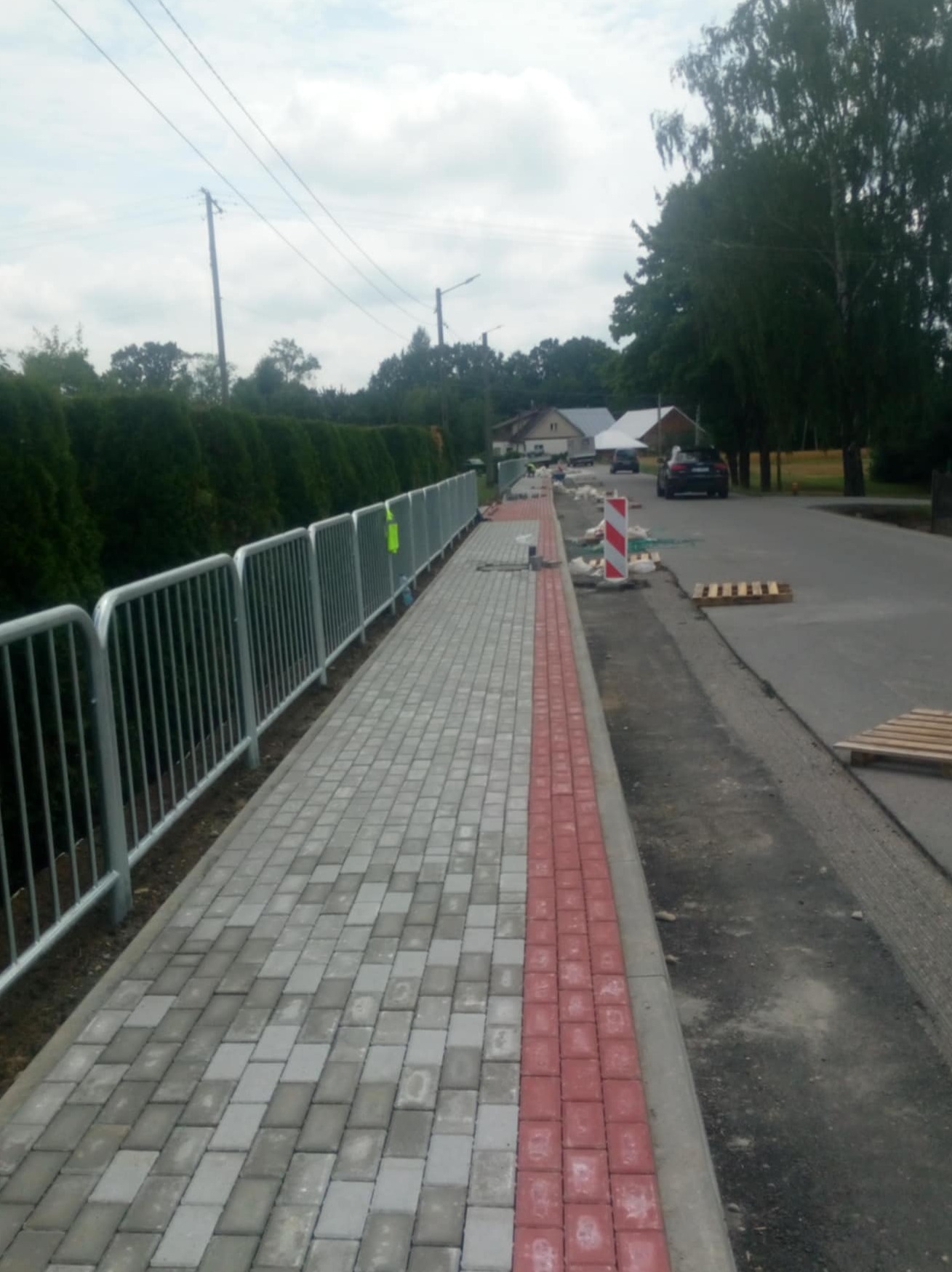 Nowo ułożony chodnik z szarej kostki brukowej z czerwonym obramowaniem, oddzielony od jezdni metalowym płotkiem, w tle domy i zieleń.