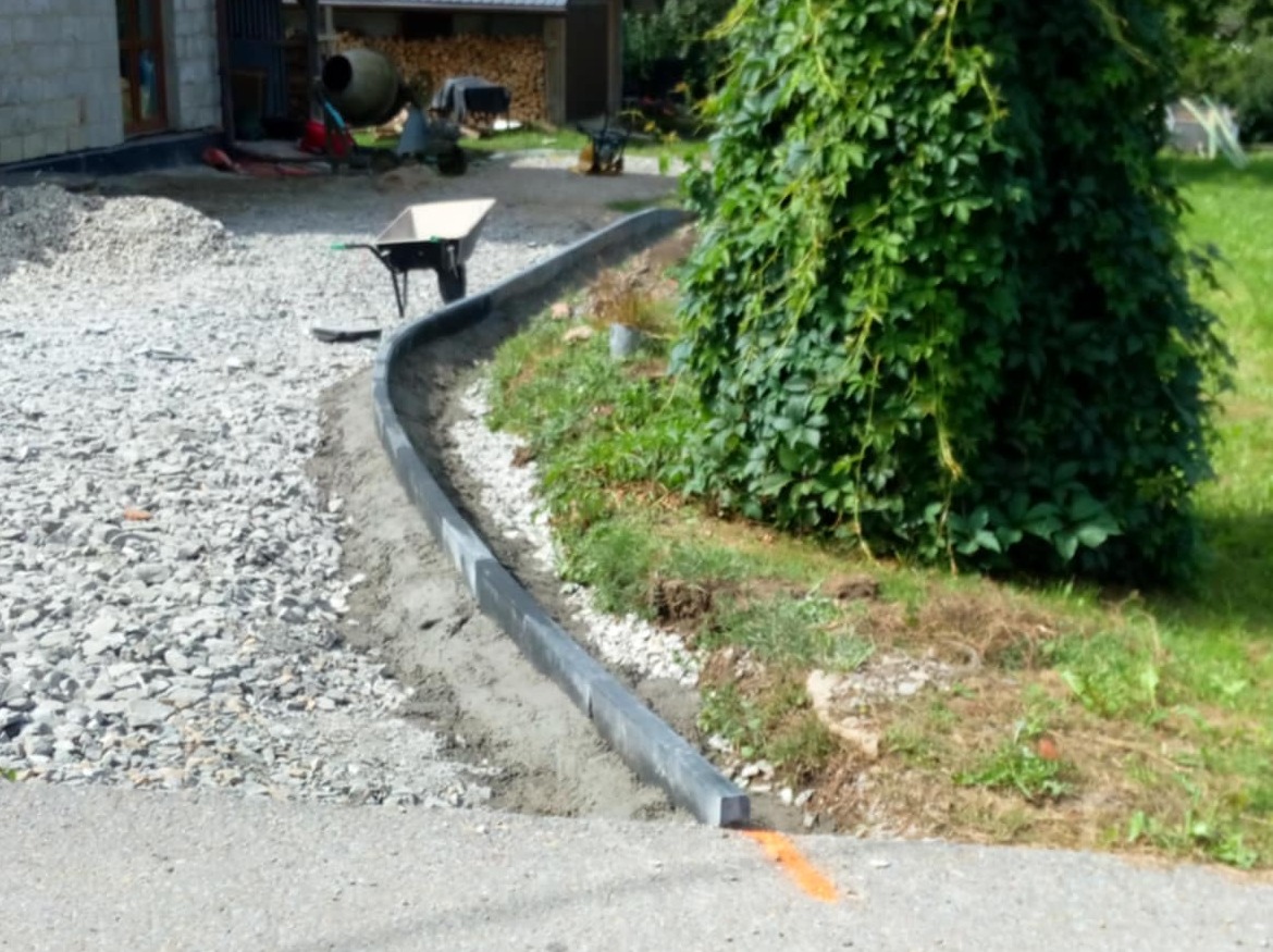 Krawężnik z betonowych elementów oddziela obszar z szarą kostką od trawnika z zieloną roślinnością, w tle widoczna taczka i betoniarka.