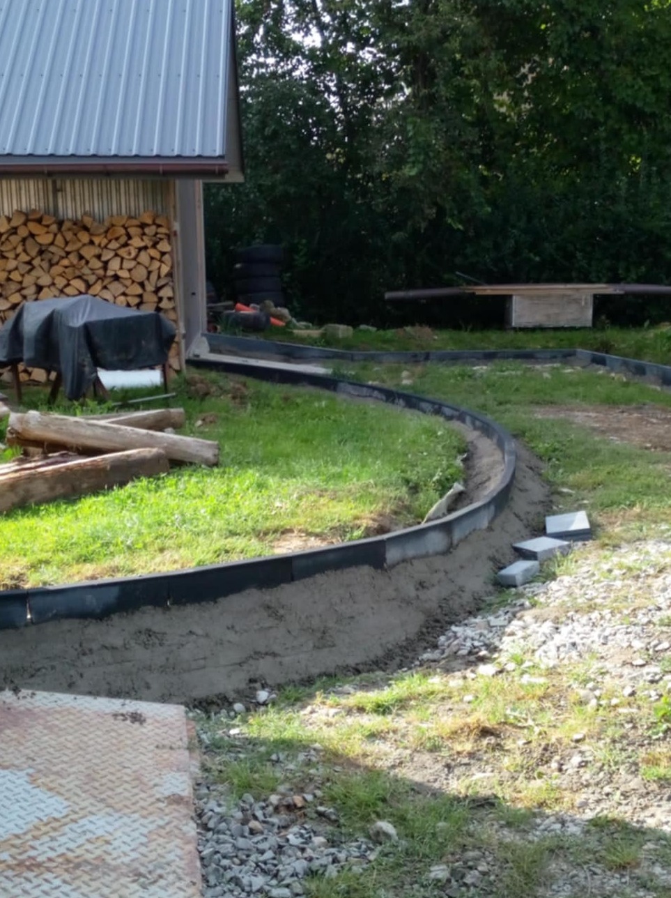 Przygotowanie podłoża pod ścieżkę z obrzeżami, widoczne ułożone krawężniki i podsypka cementowa, w tle sterta drewna i zieleń.