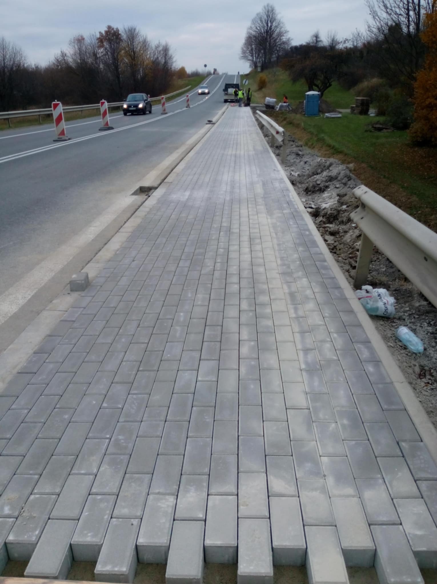 Świeżo ułożona, szara kostka brukowa tworząca chodnik wzdłuż drogi, z widocznymi samochodami w oddali i elementami zabezpieczającymi budowę.