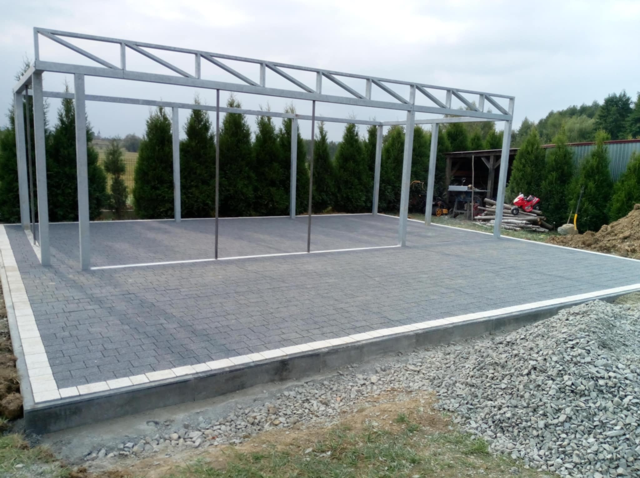 Ułożona szara kostka brukowa z jasnym obramowaniem, przygotowana pod konstrukcję z metalowych profili, w tle rząd tuj i mały budynek gospodarczy, częściowo widoczna sterta gruzu.